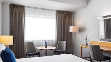 A hotel room with a large bed, two chairs, a small round table, a desk, a lamp, and a mirror.