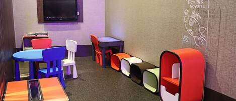 A children's play area with colorful tables and chairs, a flat-screen TV, and a wall with a family-themed drawing.