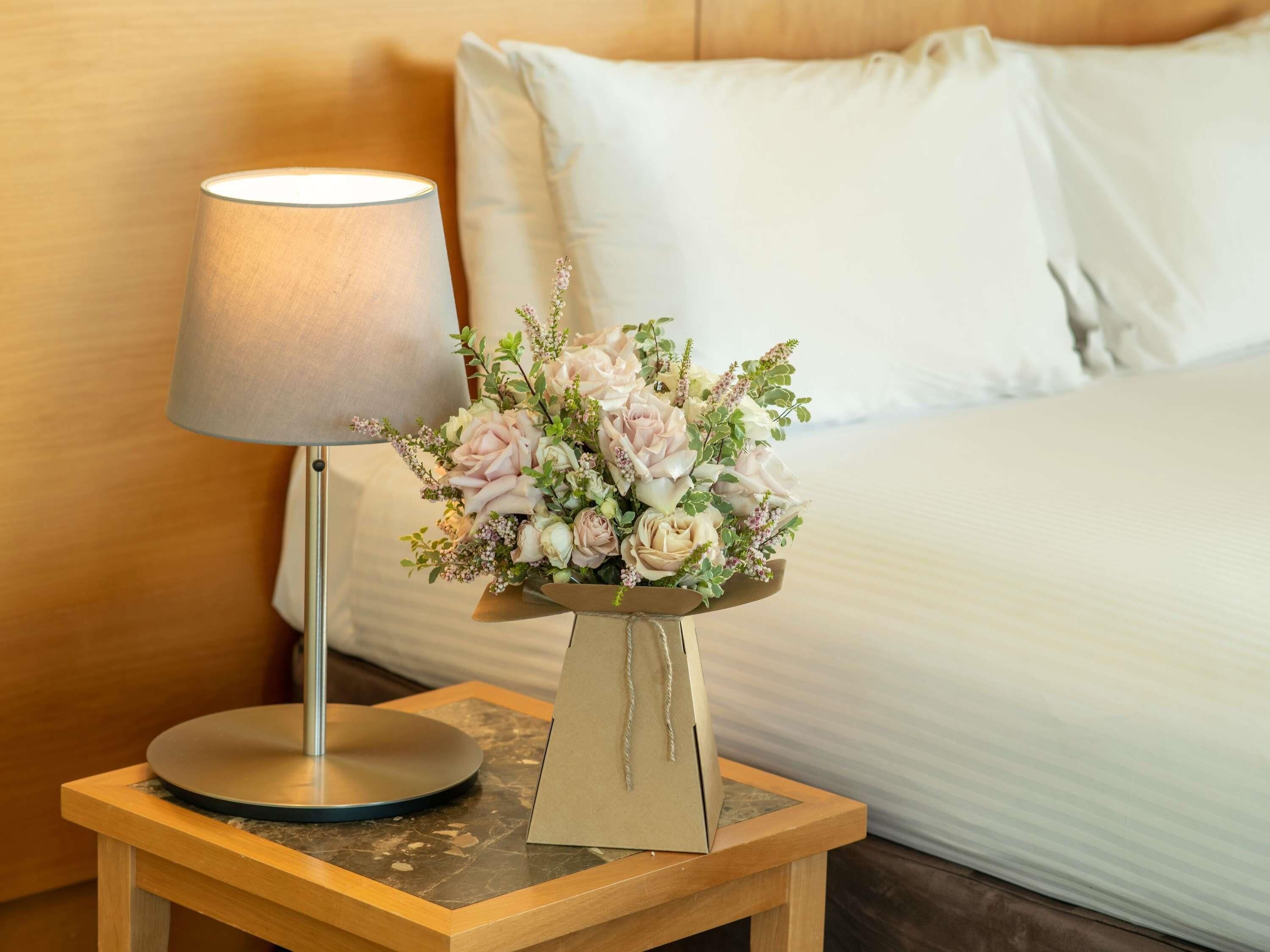 A bed with white linens, a bedside lamp, and a floral arrangement on a wooden nightstand.