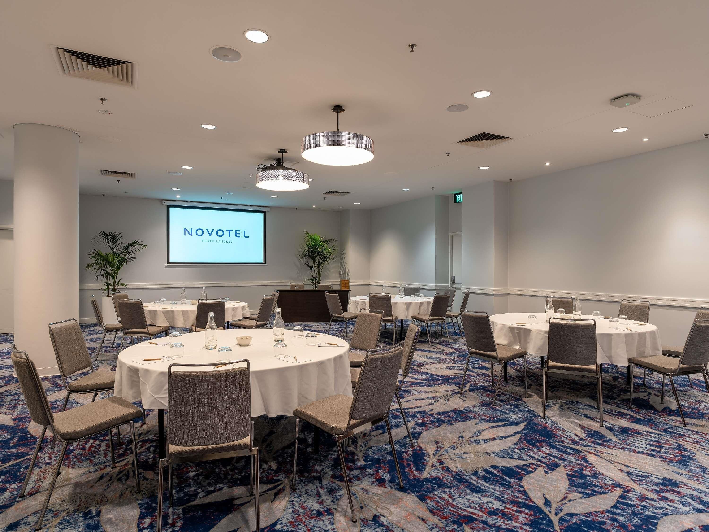 A conference room with multiple round tables, chairs, a projector screen displaying 'NOVOTEL,' and a carpeted floor.