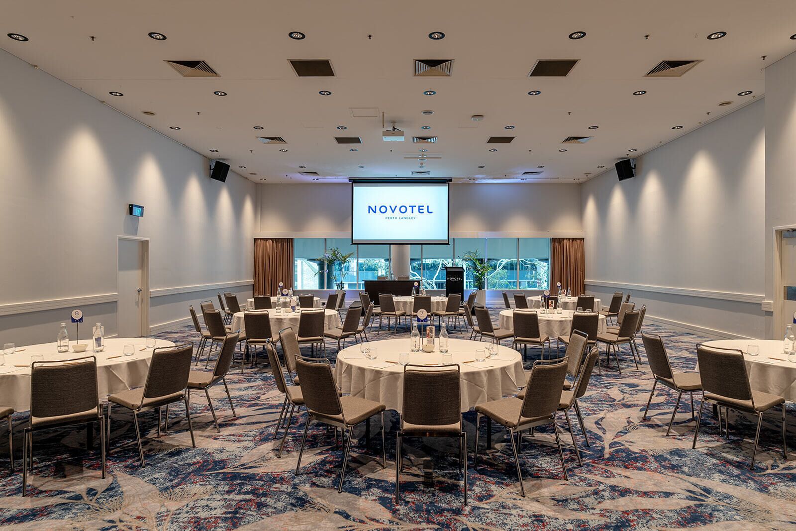A conference room with multiple round tables, chairs, and a projection screen displaying 'NOVOTEL'.