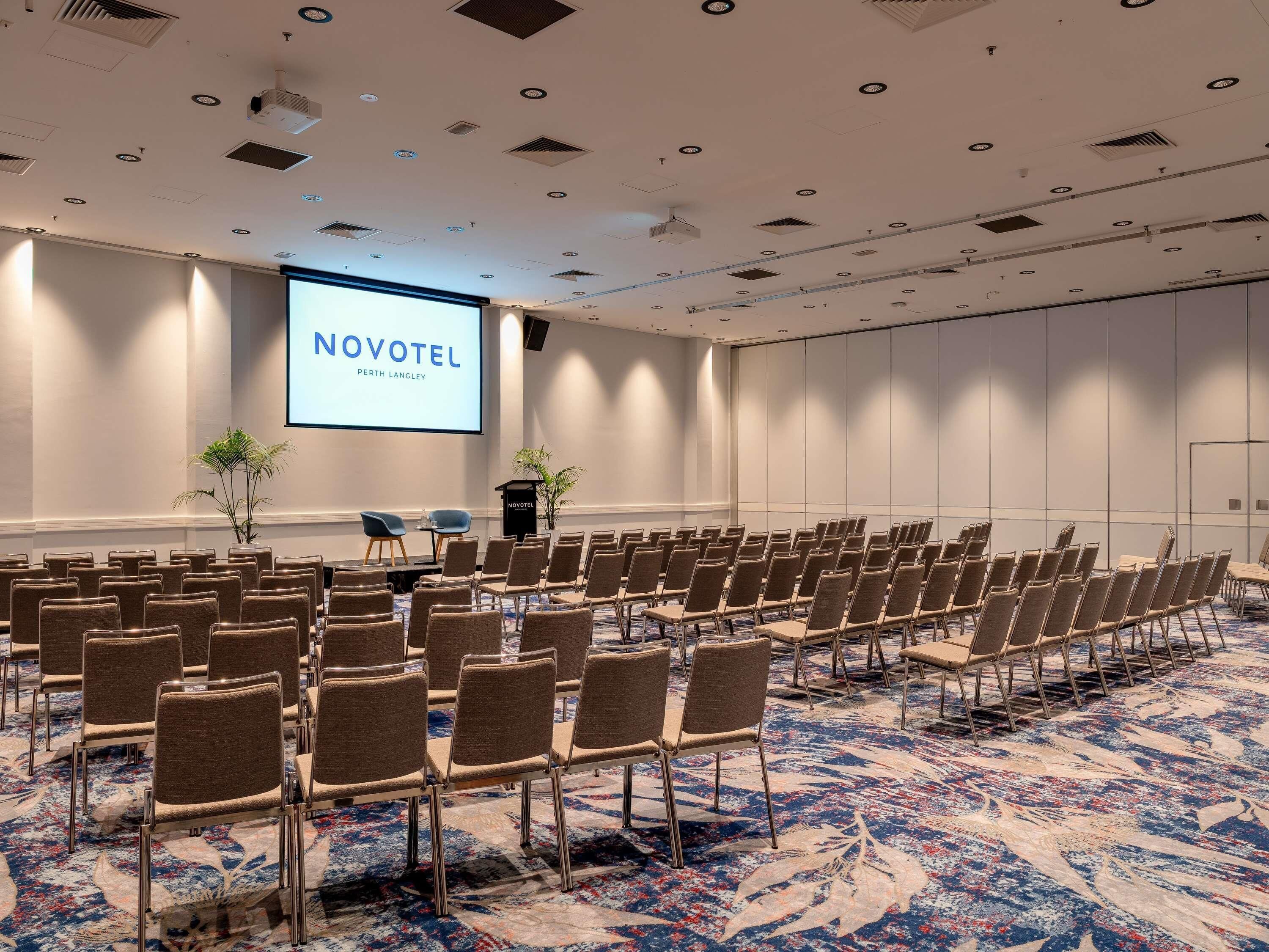 Une salle de conférence avec des rangées de chaises, un écran de projection affichant le texte « NOVOTEL PERTH LANGLEY », et un sol recouvert de moquette.