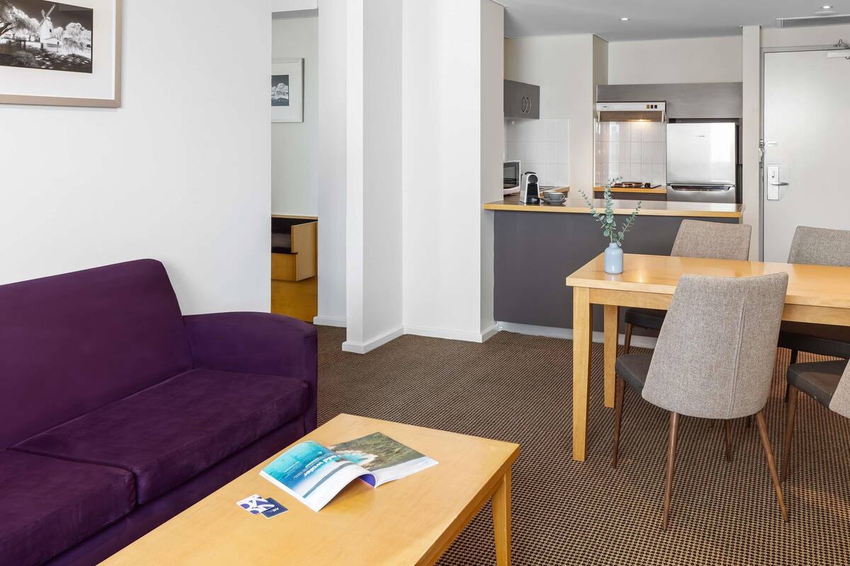 A modern hotel room with a purple sofa, a wooden table with a magazine, a dining area with a table and chairs, and a kitchenette in the background.