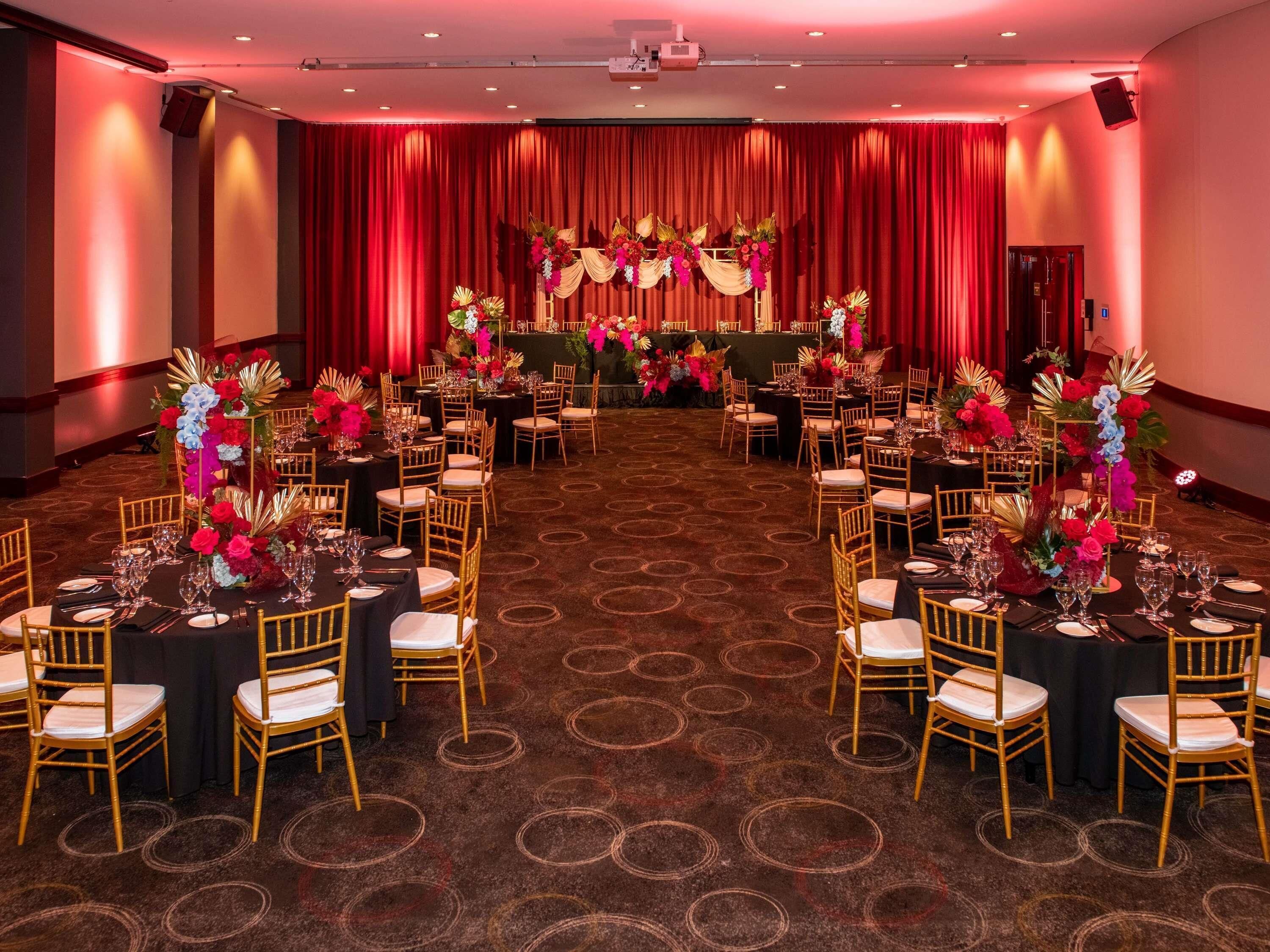 A banquet hall with round tables, gold chairs, and red curtains.