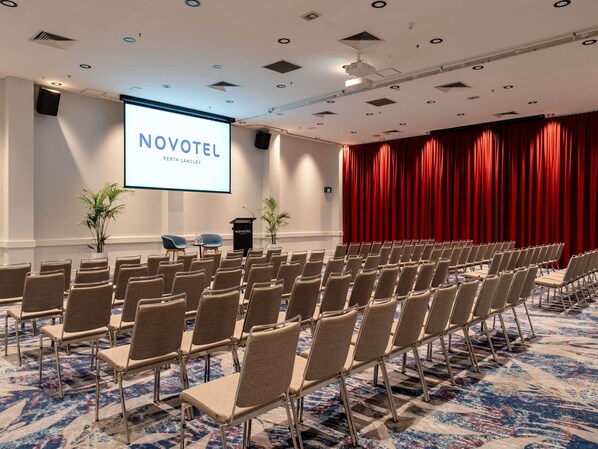 A conference room with rows of chairs, a podium, and a projection screen displaying 'NOVOTEL PERTH LANGLEY'.