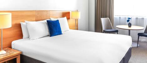 Una habitación de hotel con una cama grande, dos lámparas de noche, una mesita redonda con una silla y una ventana con cortinas.