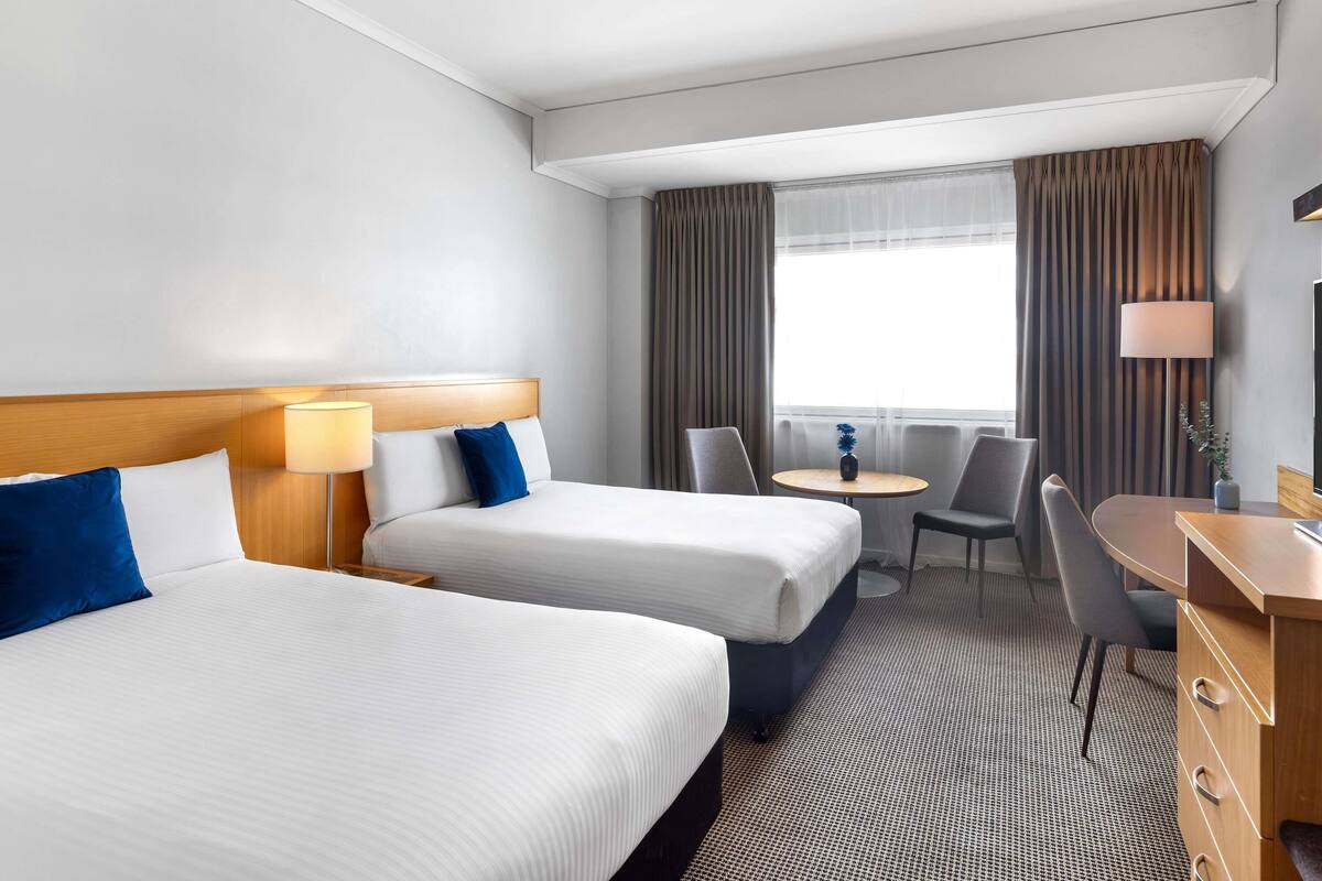 A hotel room with two beds, a desk, a chair, a small table, and a TV.