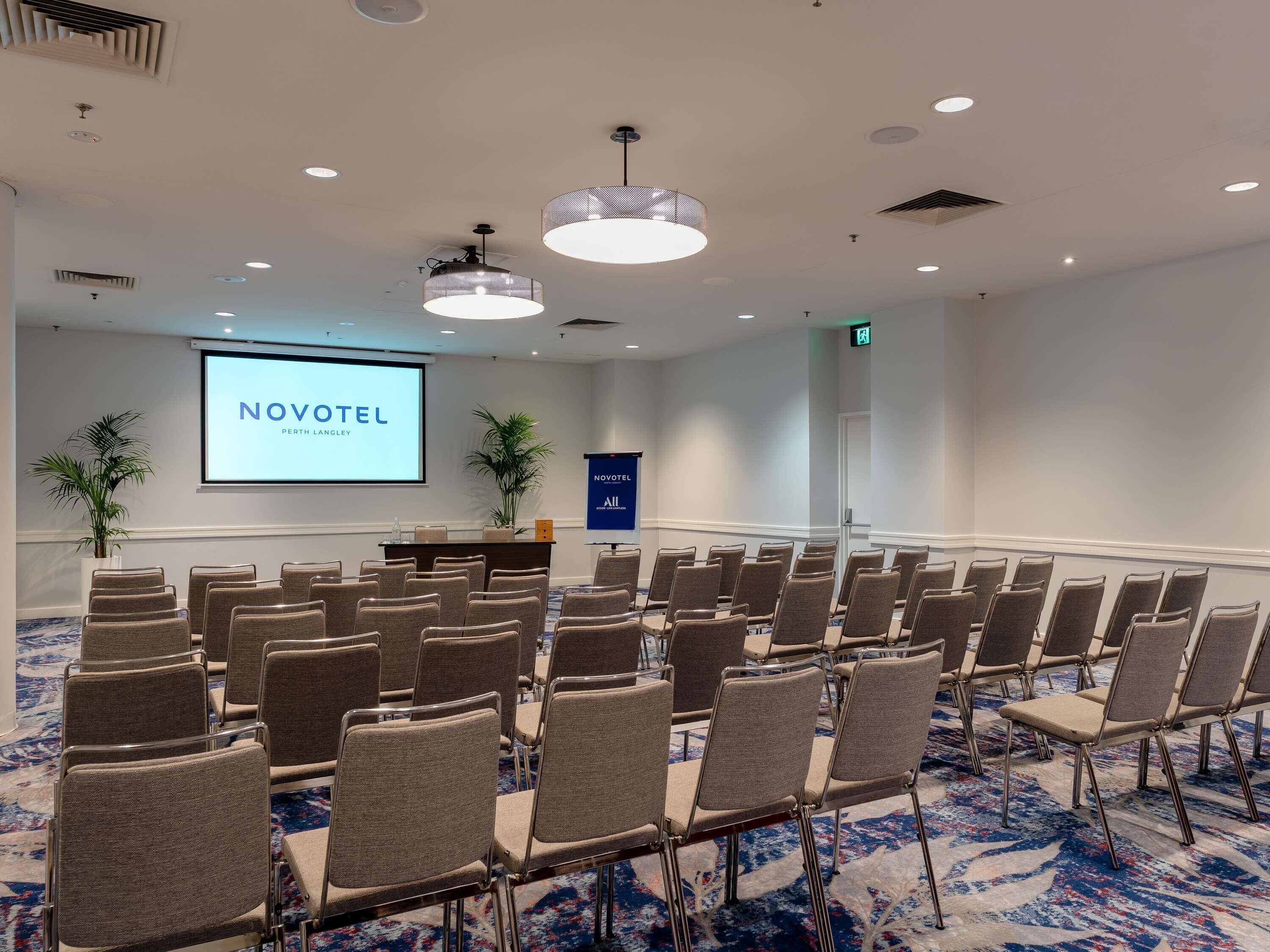 Un salón de conferencias con filas de sillas, un atril, una pantalla que muestra 'NOVOTEL PERTH LANGLEY' y una alfombra azul.