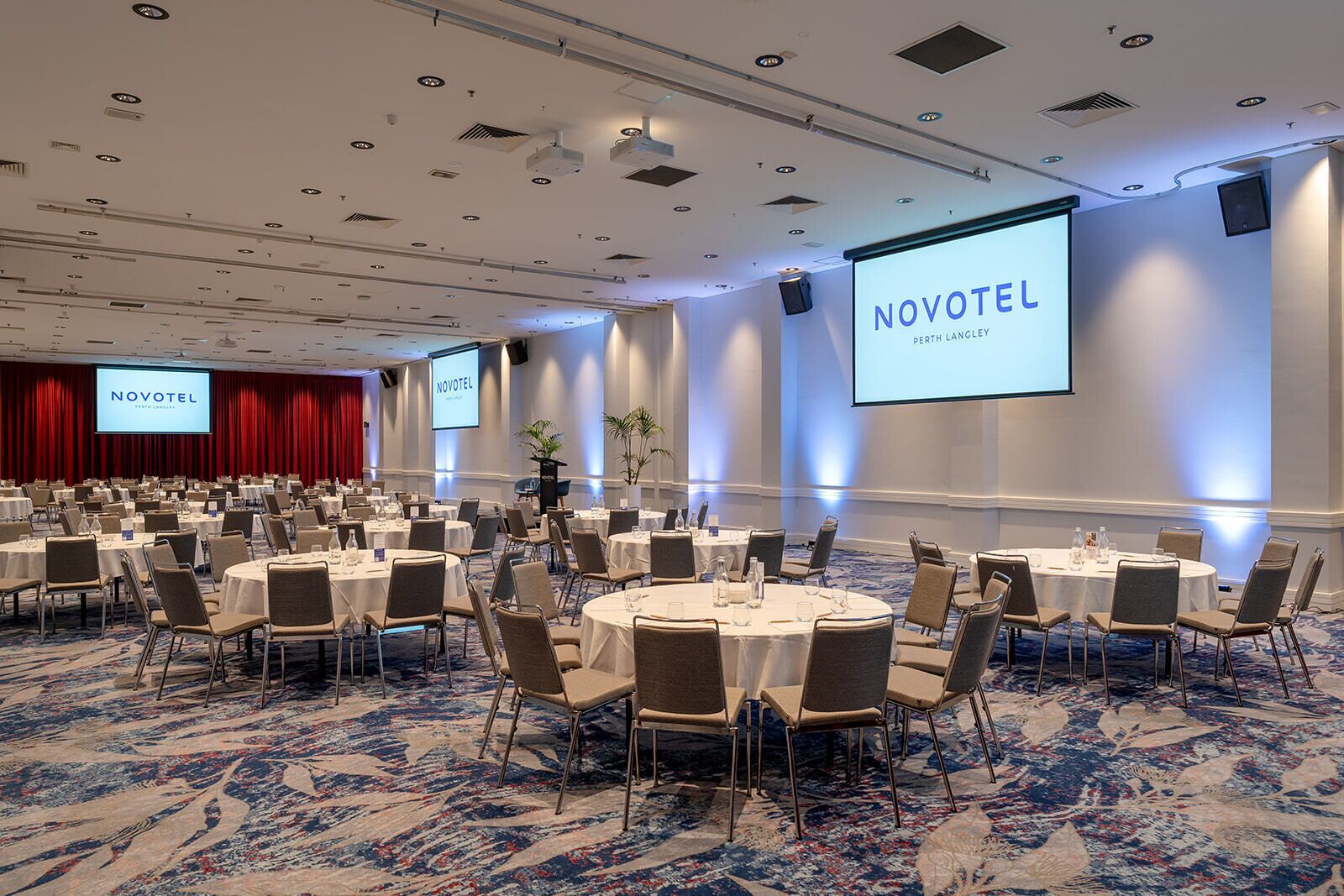 A conference room with multiple tables and chairs, a large screen displaying 'NOVOTEL Perth Langley', and a carpeted floor.