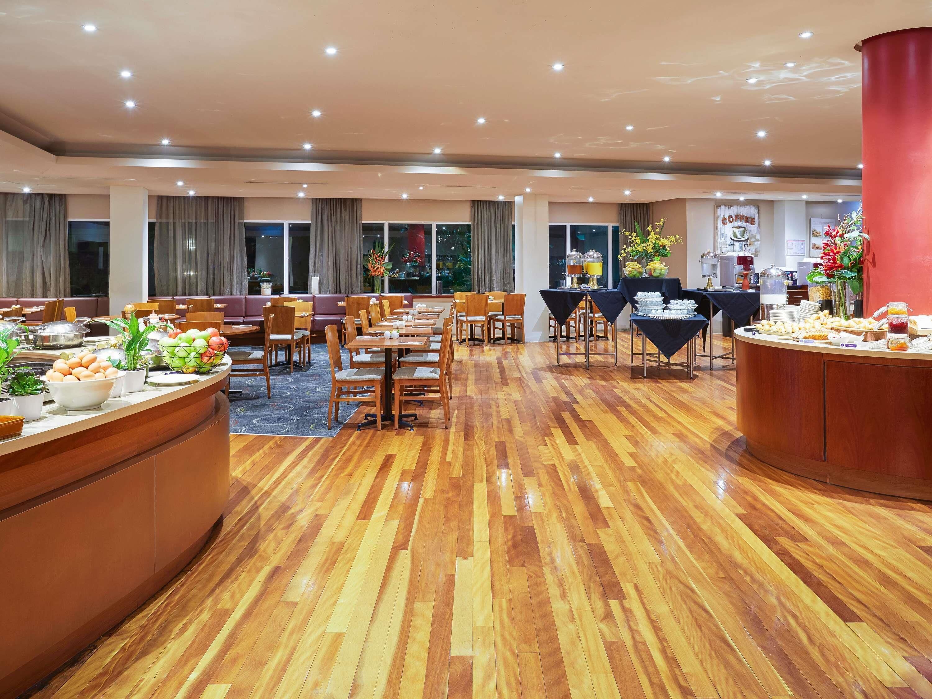 A spacious dining area with multiple tables and chairs, a buffet counter with food and drinks, and large windows providing natural light.