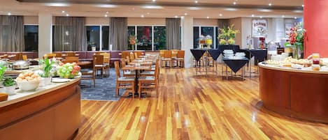 A spacious dining area with multiple tables and chairs, a buffet counter with food and drinks, and large windows providing natural light.