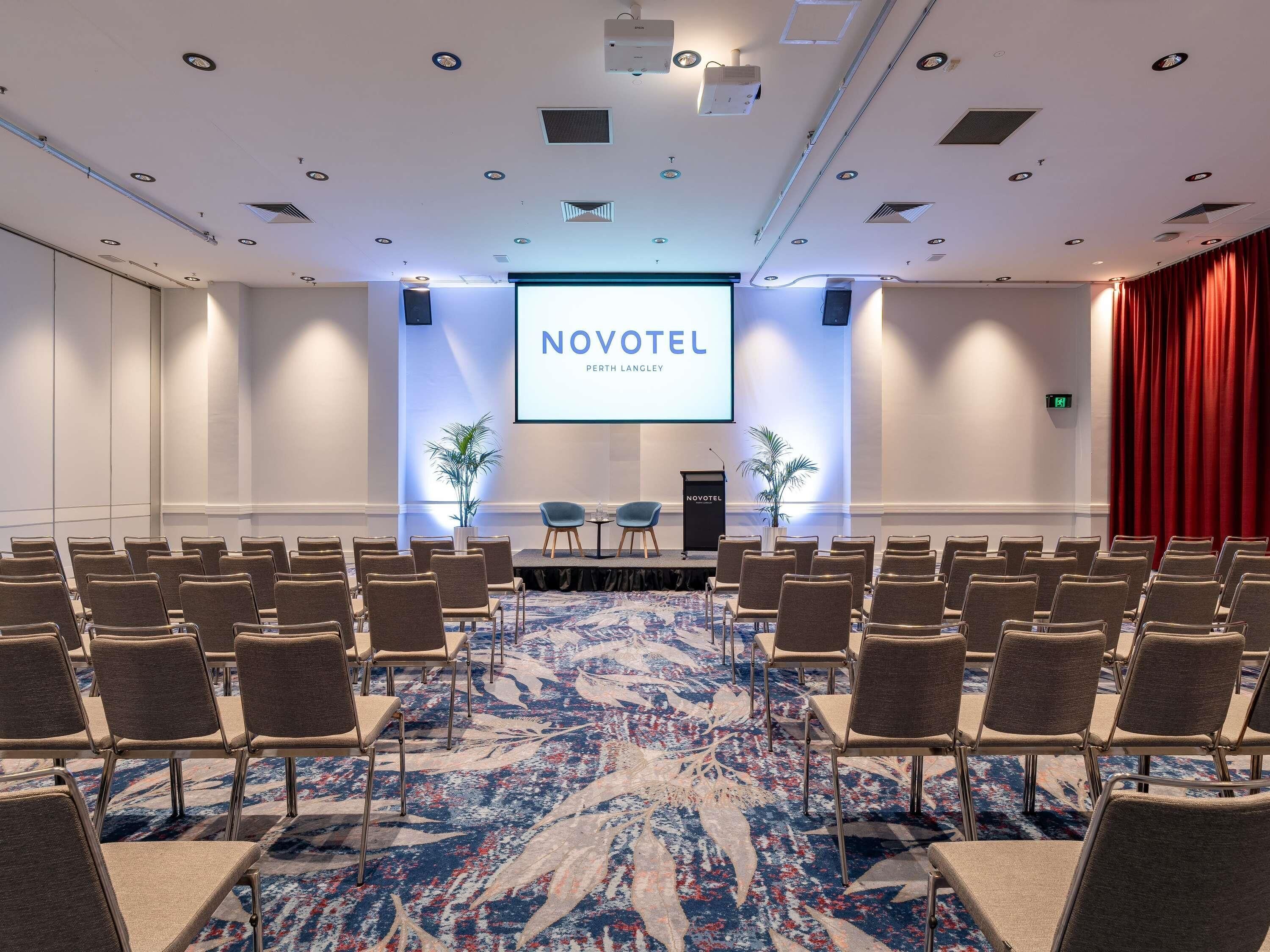 Uma sala de conferências com fileiras de cadeiras, um pódio e uma tela exibindo 'NOVOTEL PERTH LANGLEY'.
