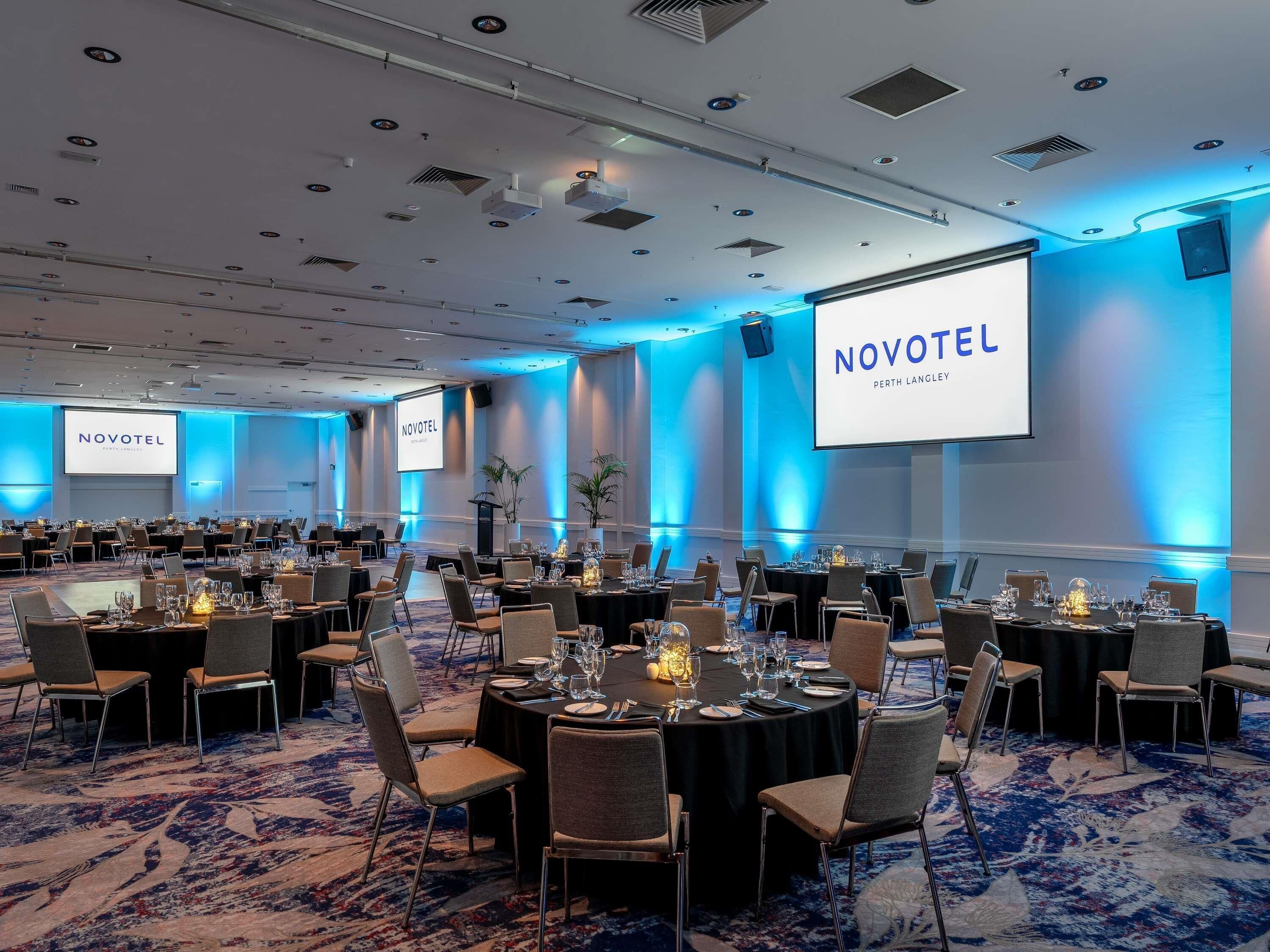 A conference room at Novotel Perth Langley with multiple tables set for meetings, a large screen displaying the hotel's name, and a carpeted floor.