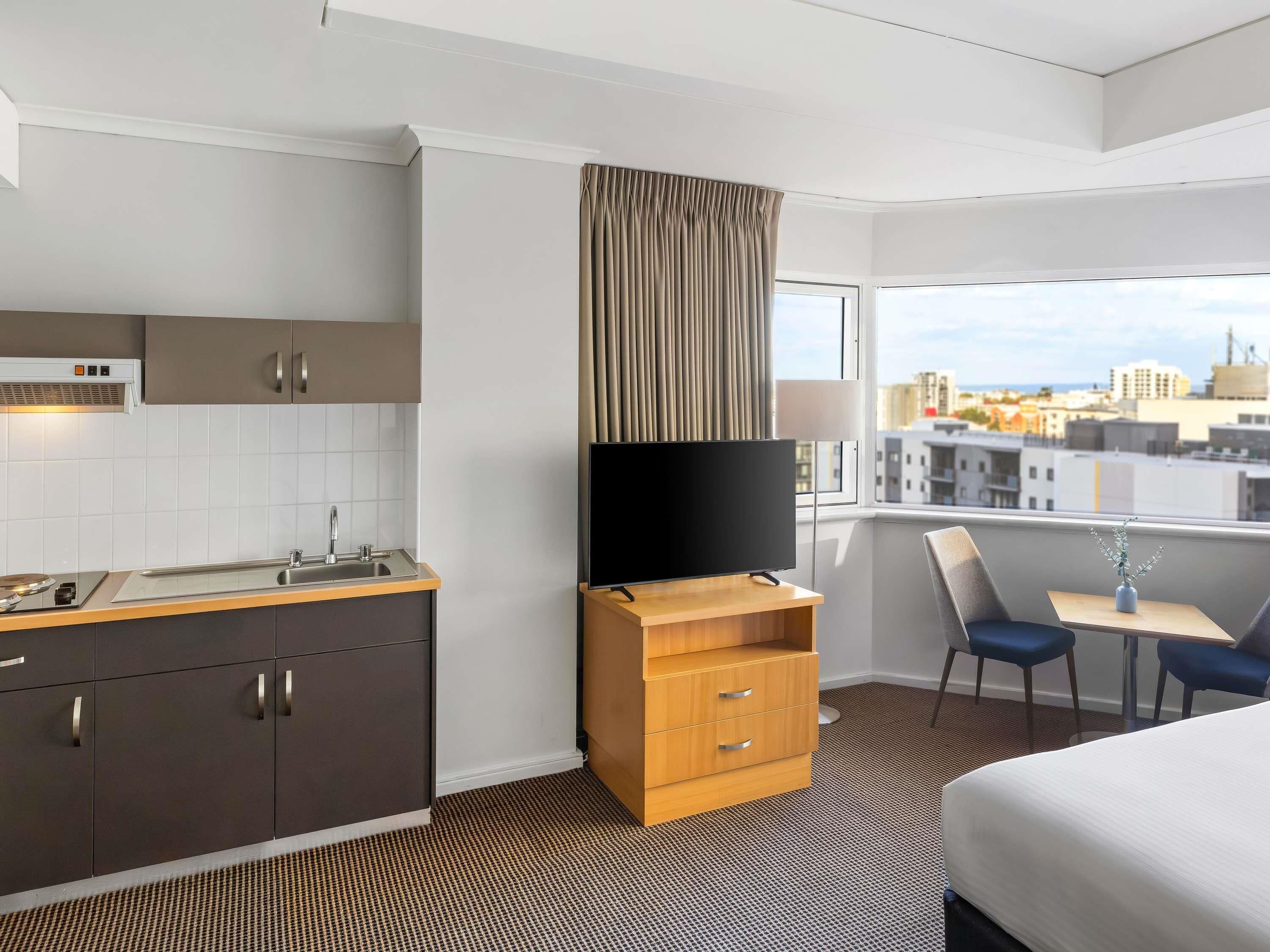 Una habitación de hotel moderna con kitchenette, televisor de pantalla plana, comedor con mesa y sillas, y un ventanal con vistas a la ciudad.