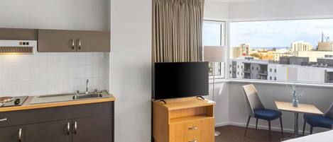 Una habitación de hotel moderna con kitchenette, televisor de pantalla plana, comedor con mesa y sillas, y un ventanal con vistas a la ciudad.