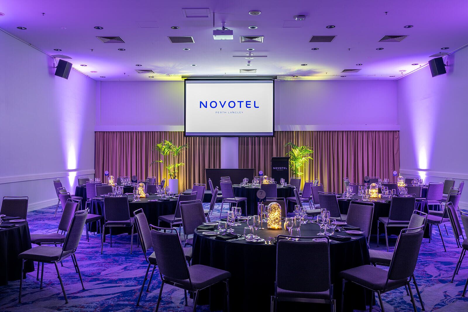 A conference room at Novotel Perth Langley with multiple tables, chairs, and a large screen displaying the hotel name.