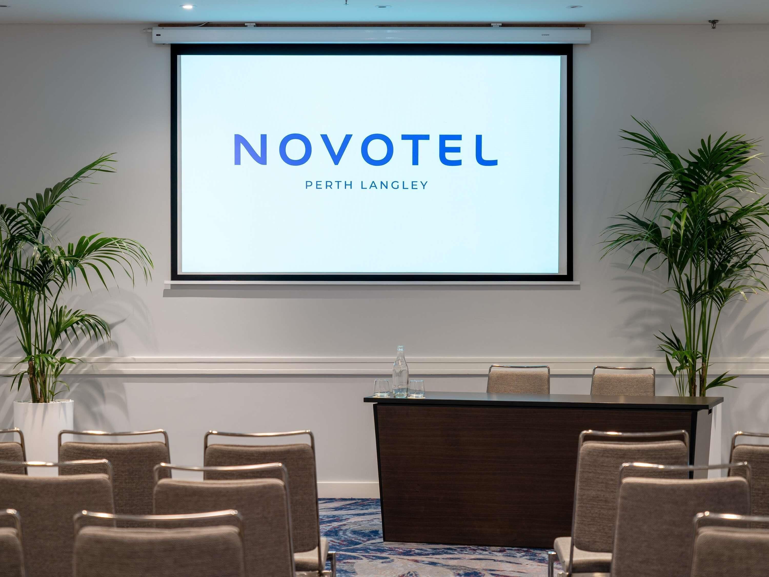 A conference room with a screen displaying 'NOVOTEL PERTH LANGLEY,' a podium, and several chairs.