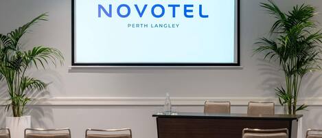 A conference room with a screen displaying 'NOVOTEL PERTH LANGLEY,' a podium, and several chairs.