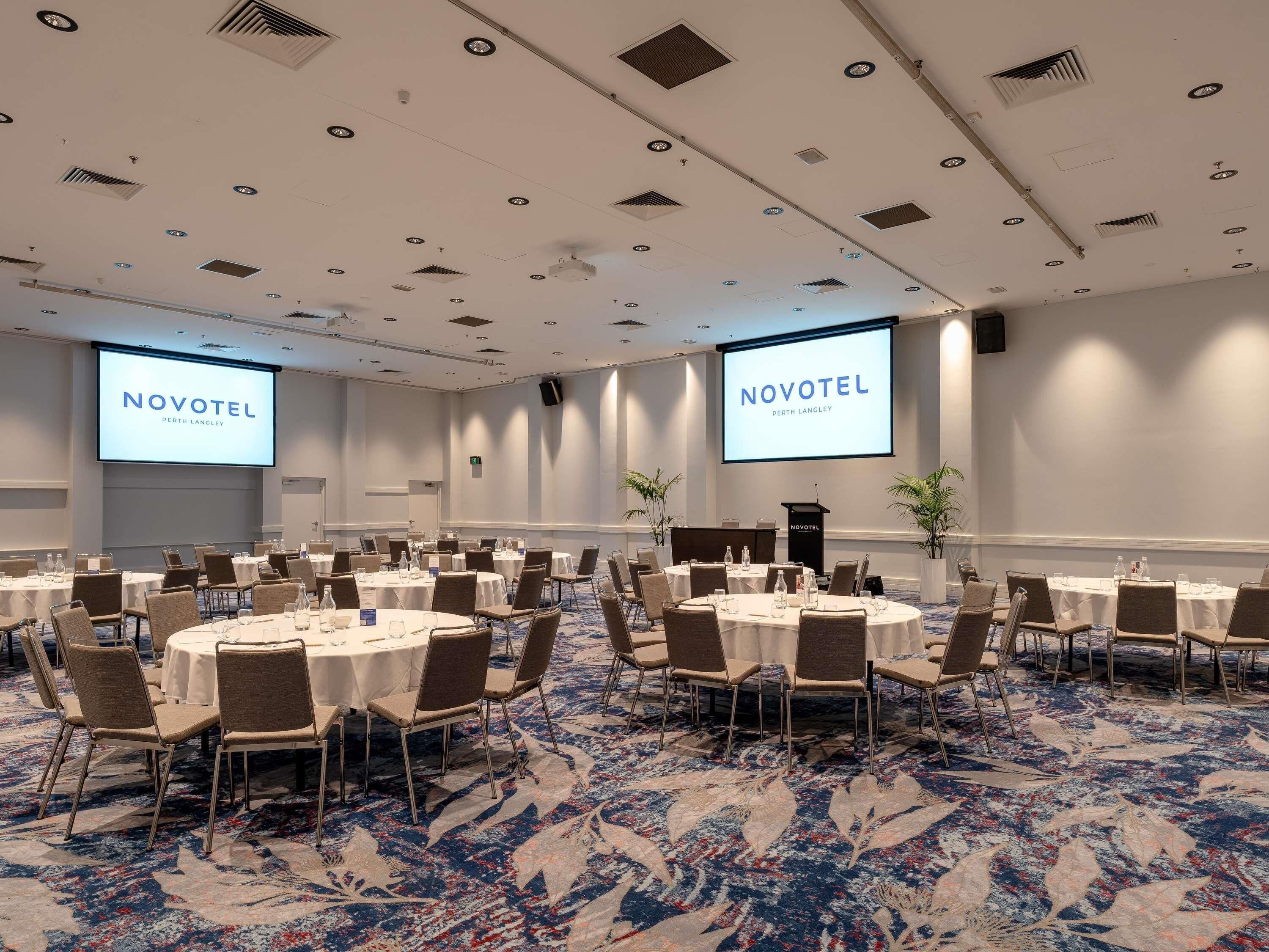 A conference room with multiple round tables, chairs, and a large screen displaying 'NOVOTEL PERTH LANGLEY'.