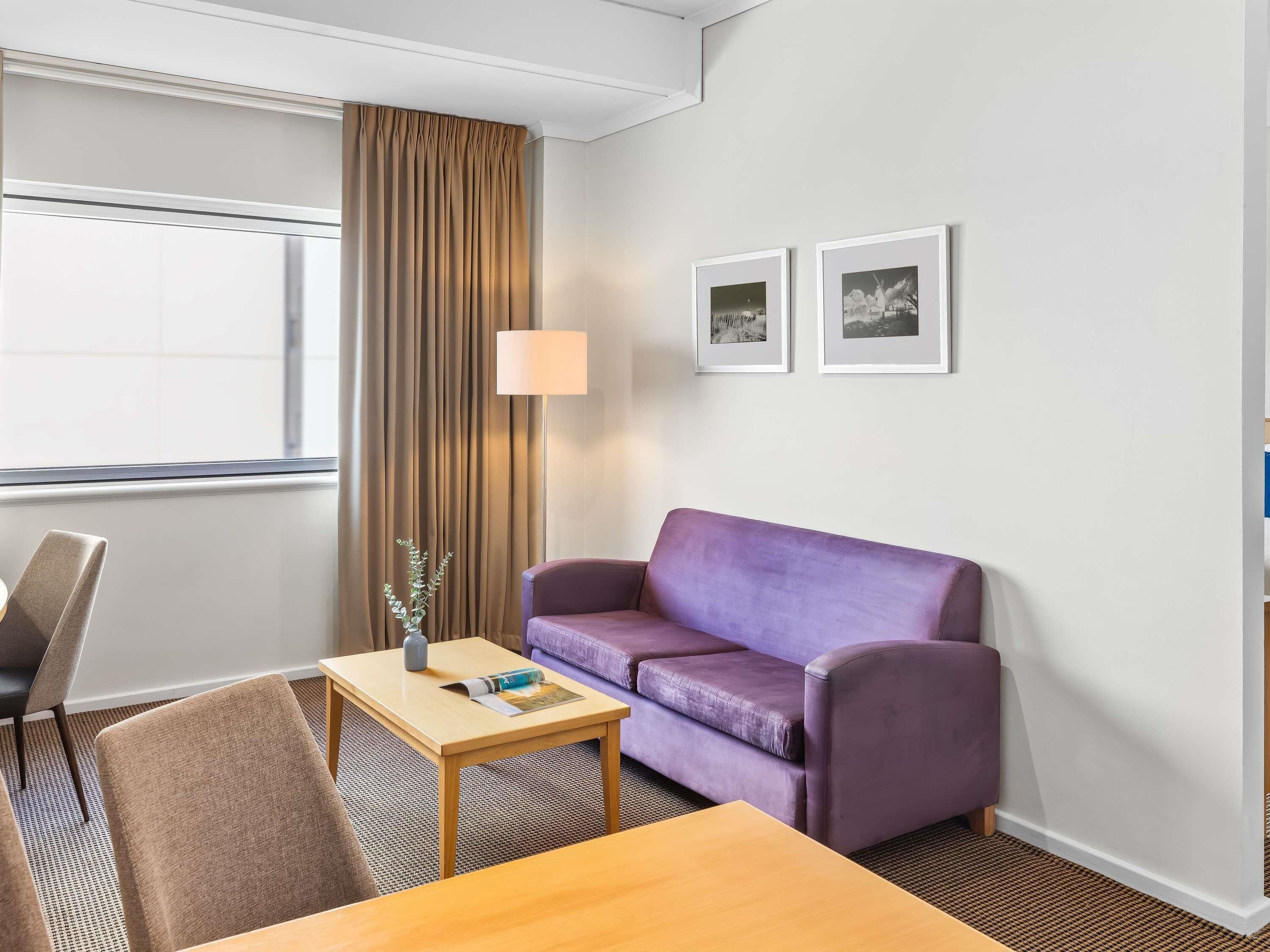 A hotel room with a purple sofa, a wooden table, a lamp, and two framed pictures on the wall.