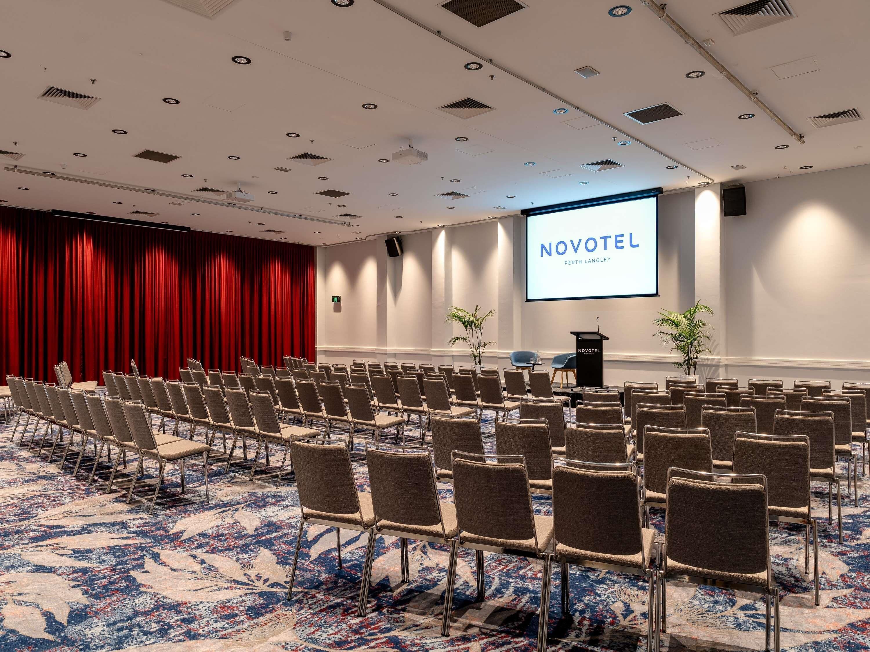 Un salón de conferencias con filas de sillas, un atril y una pantalla mostrando 'NOVOTEL PERTH LANGLEY'.