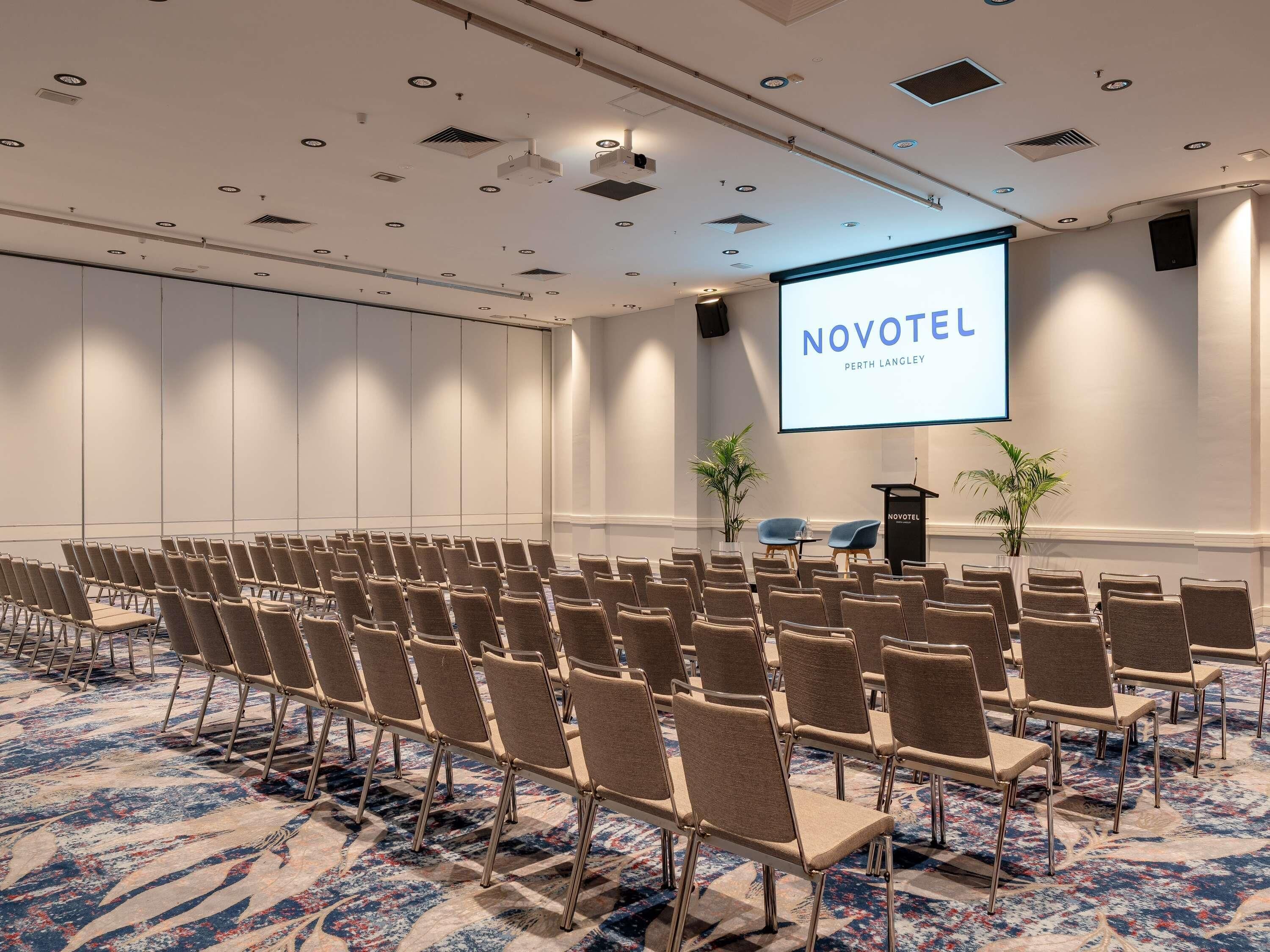 A conference room with rows of chairs, a podium, and a screen displaying 'NOVOTEL PERTH LANGLEY'.