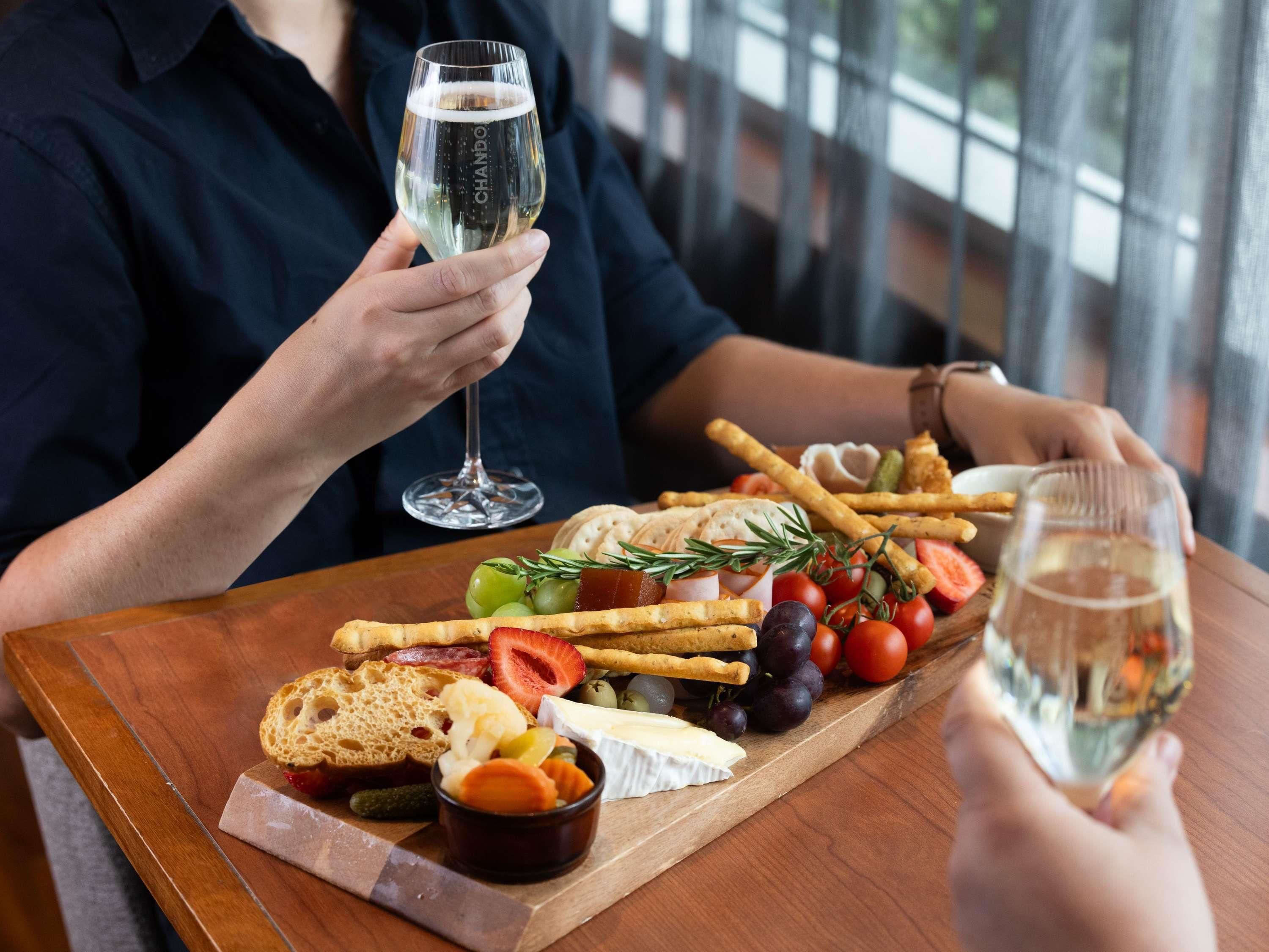 A person holding a glass of white wine, seated at a table with a variety of cheeses, fruits, and crackers.