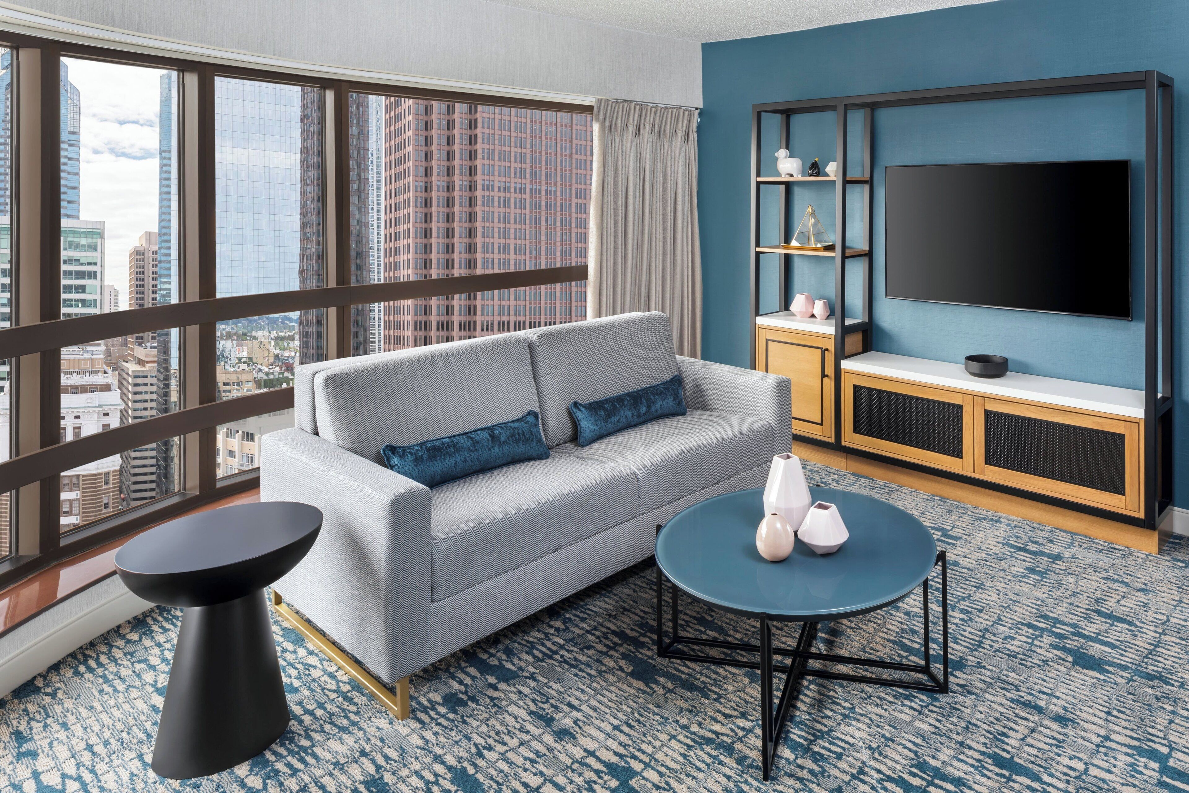A modern hotel room with a grey sofa, a round coffee table, a TV mounted on a blue wall, and a city view through large windows.