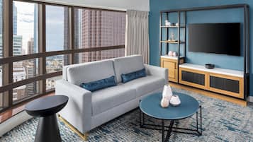 A modern hotel room with a grey sofa, a round coffee table, a TV mounted on a blue wall, and a city view through large windows.