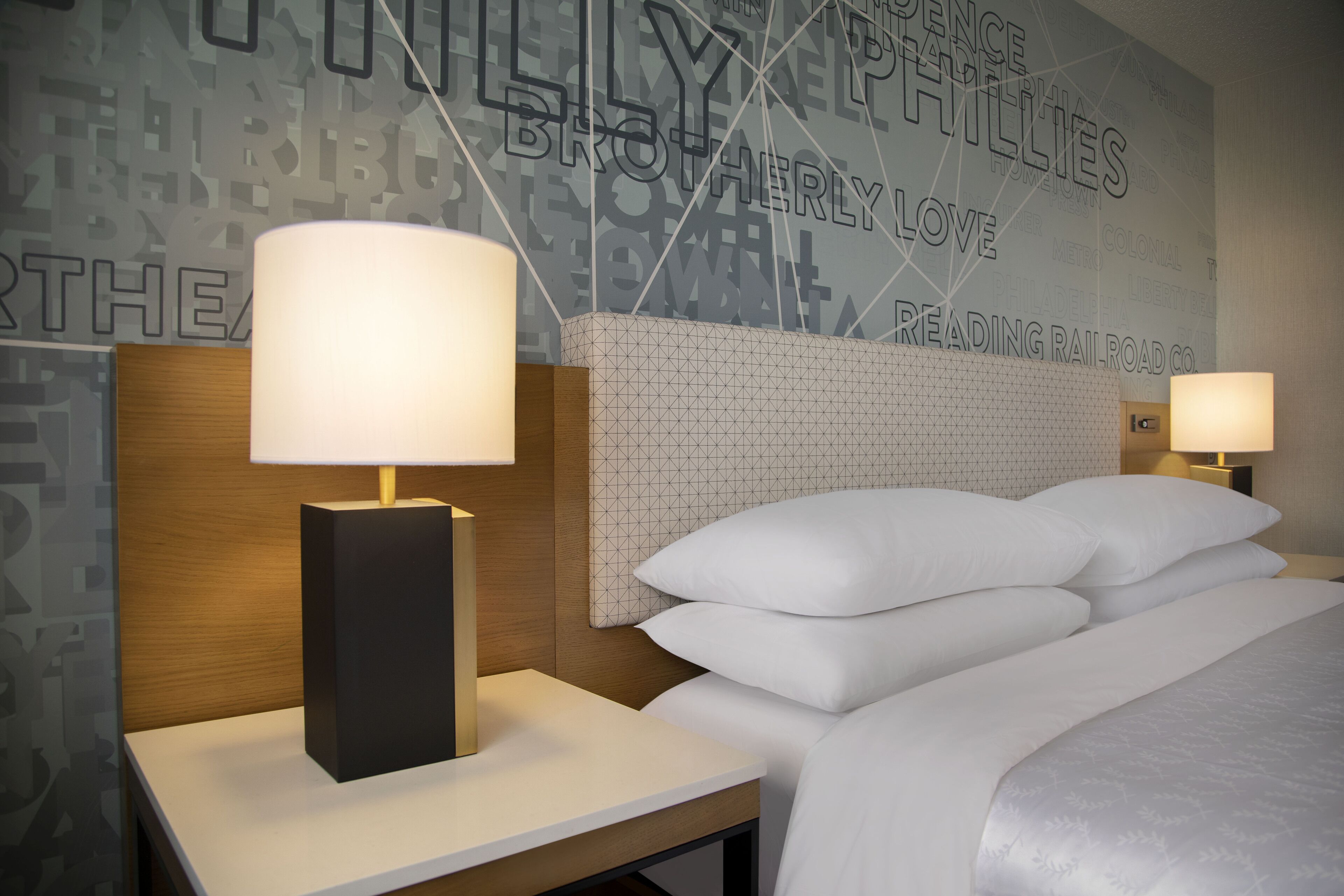 Habitación de hotel con una cama bien hecha, dos lámparas de noche y una pared con un diseño geométrico y texto.