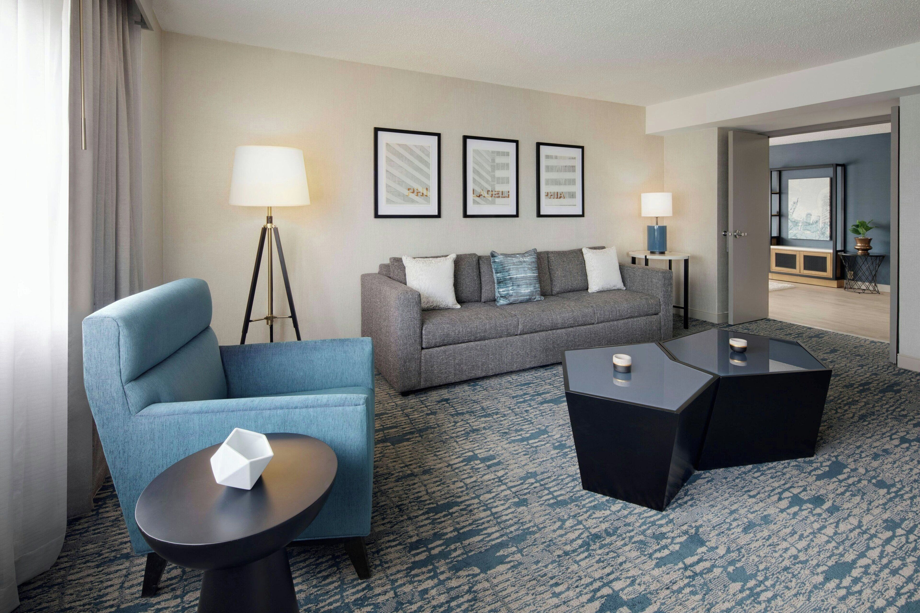 Una habitación de hotel moderna con un sillón azul, sofá gris y dos mesitas de noche negras. Hay cuadros en la pared y una lámpara de pie.