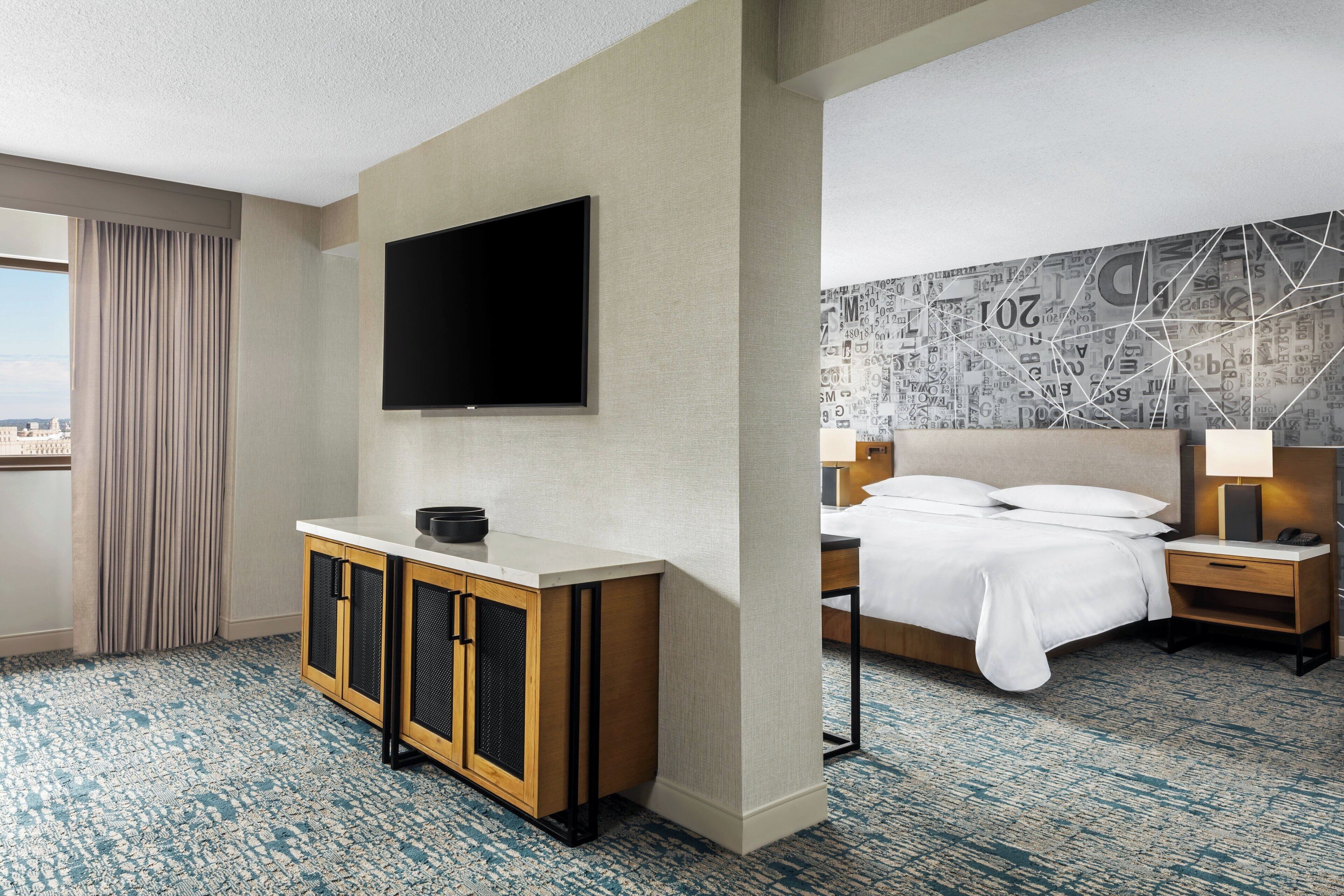 Habitación de hotel con una cama grande, televisión montada en la pared, minibar y vistas a la ciudad.