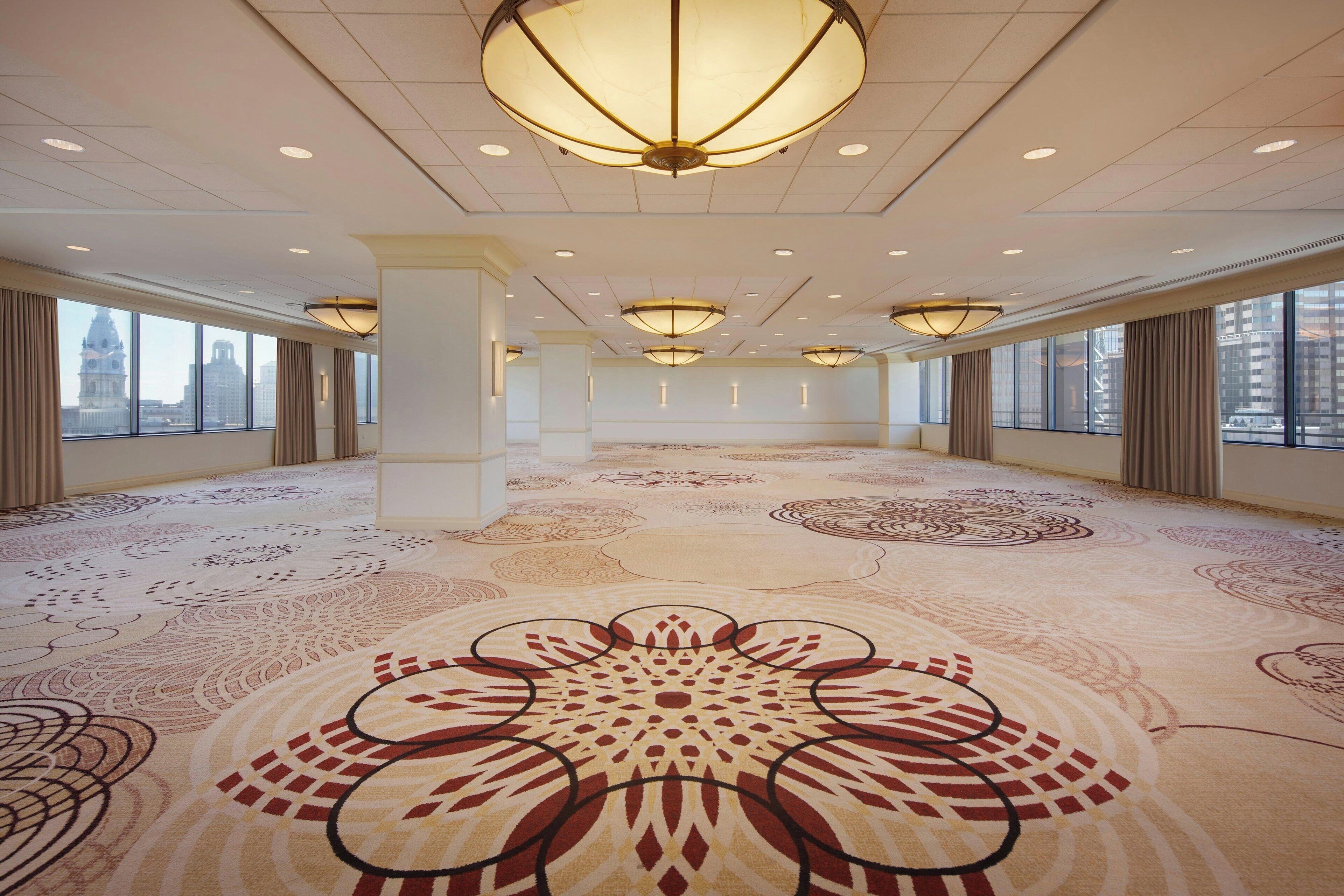 A spacious hotel lobby with large windows, patterned carpet, and modern chandeliers.