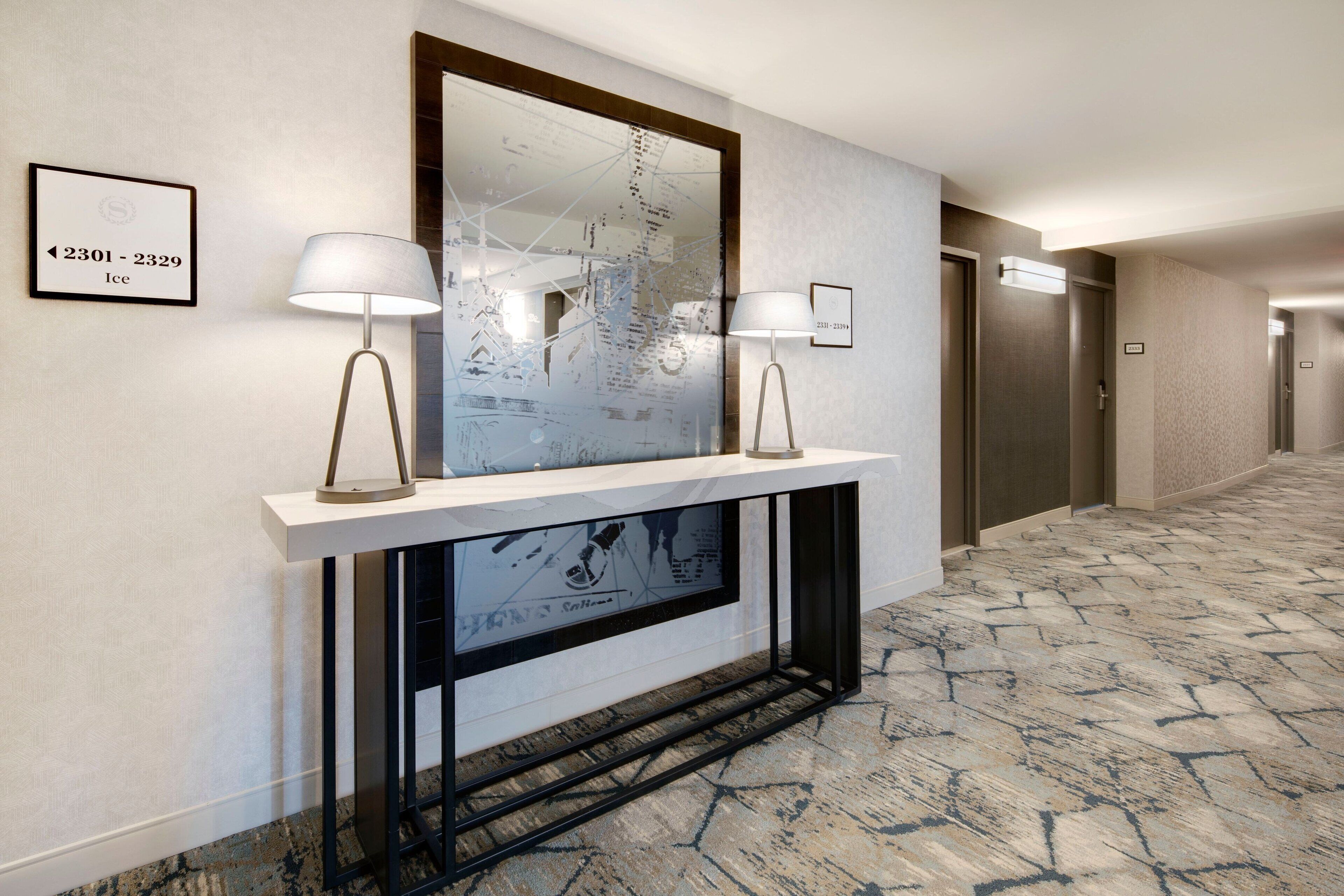 A hallway with a marble-patterned floor, a large mirror above a fireplace, and two wall-mounted lamps.