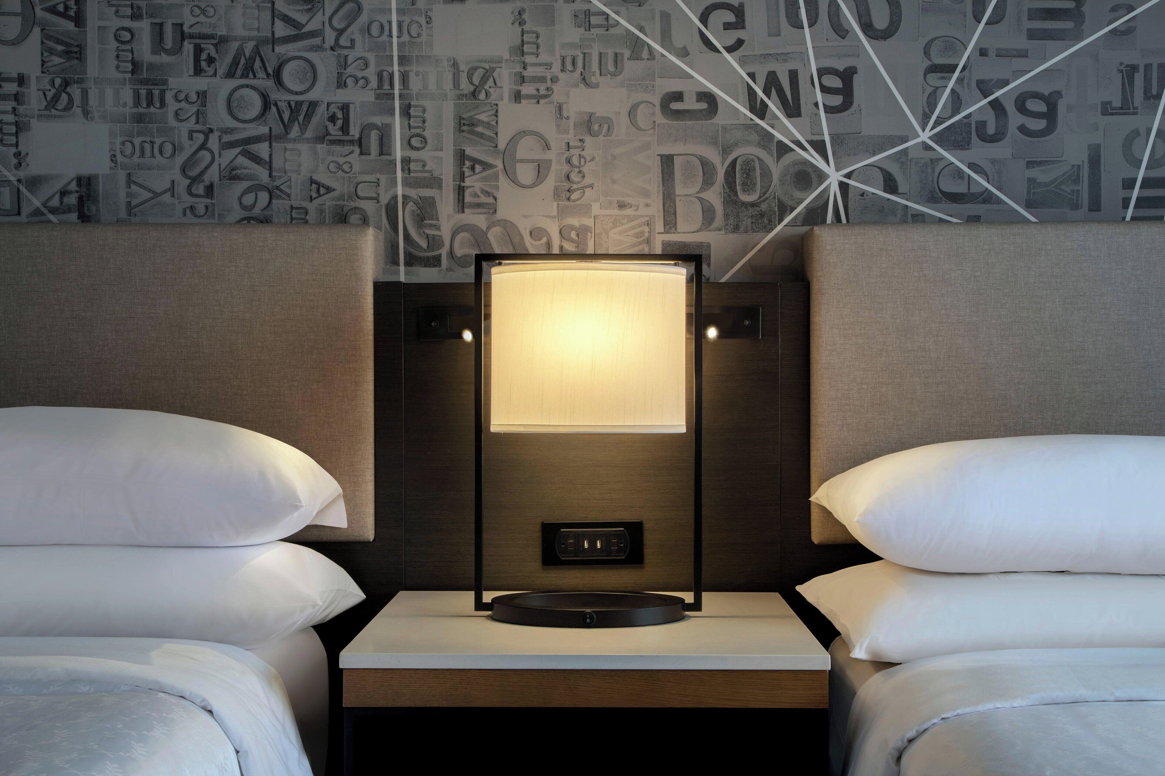 Una cama bien hecha con ropa blanca, una mesita de noche con una lámpara y una pared con diseño geométrico.