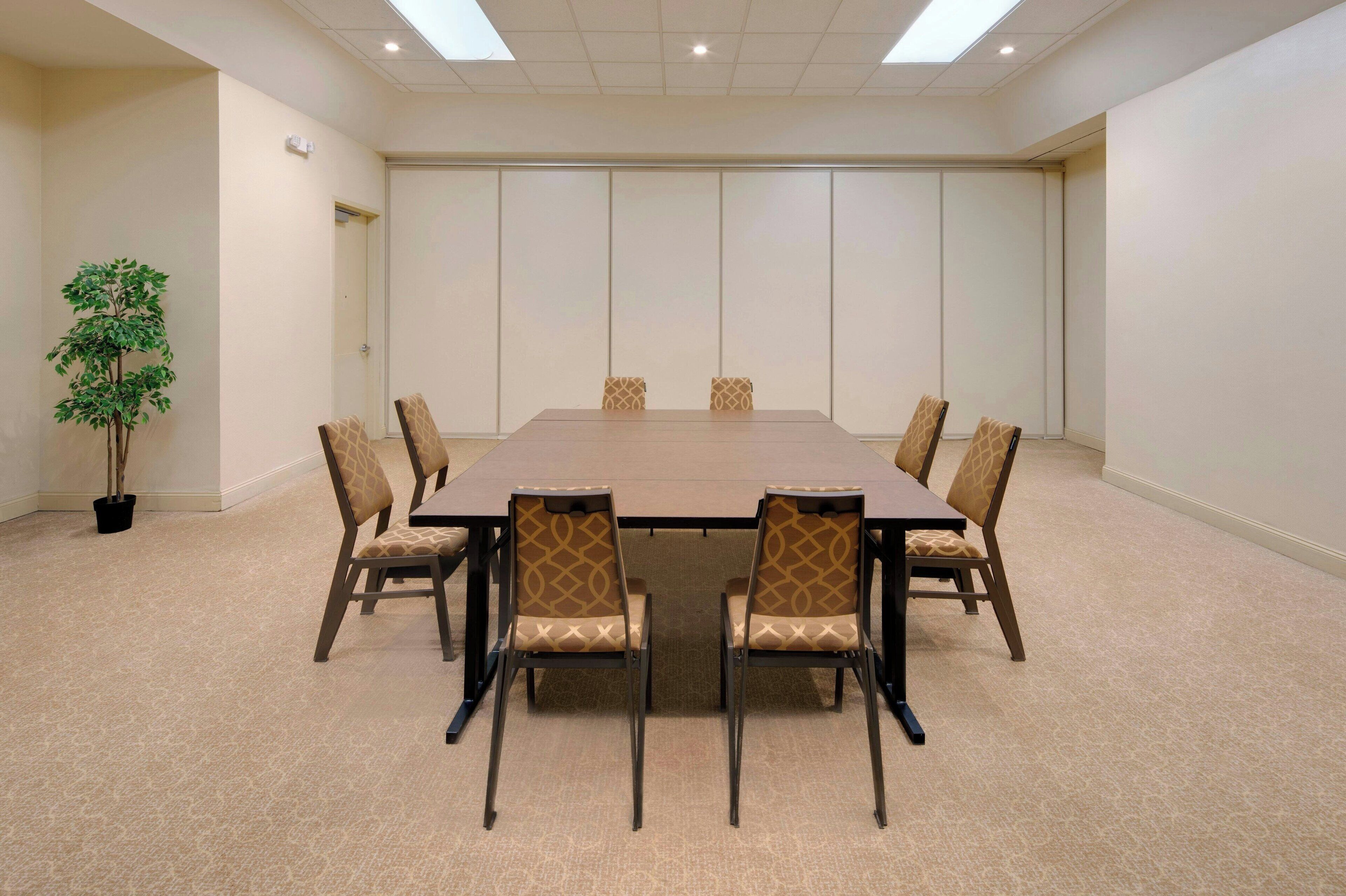 Una sala de conferencias con una gran mesa rectangular, seis sillas con tapicería estampada, una planta en maceta y una puerta al fondo.