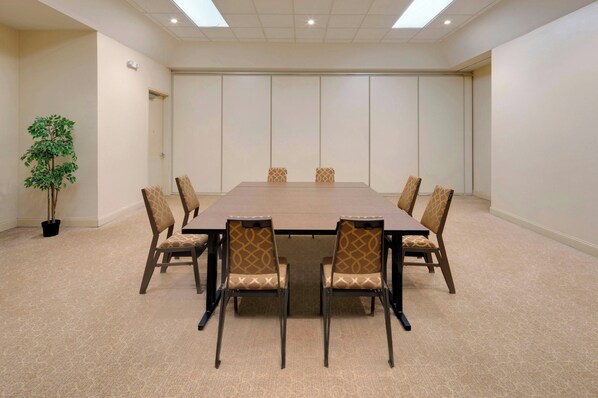 Une salle de conférence avec une grande table rectangulaire, six chaises recouvertes d’un tissu à motifs, une plante en pot et une porte visible au fond.