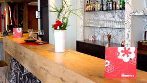 A bar area with a wooden counter, a white vase with red flowers, and shelves stocked with bottles and glasses.