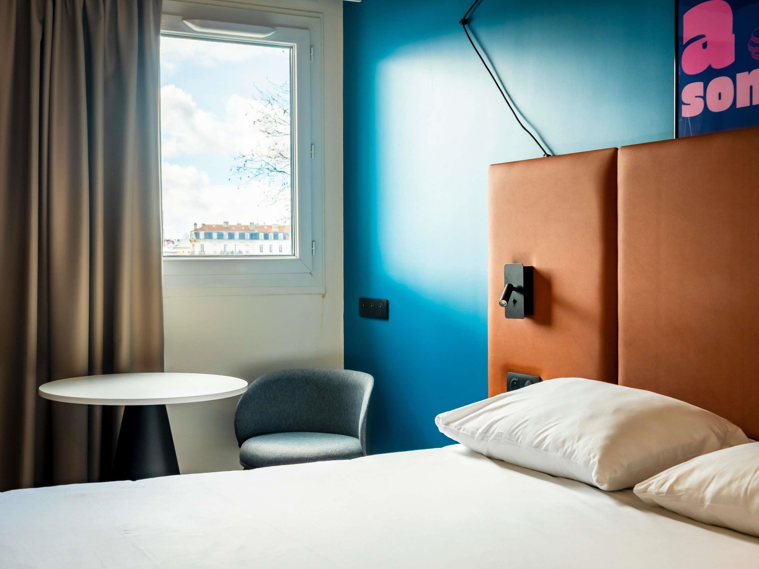 A hotel room with a bed, a small round table, a chair, a window with curtains, and a blue wall.