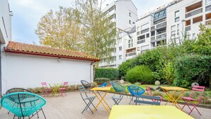 An outdoor patio with colorful chairs and tables, surrounded by greenery and a white building with a red-tiled roof. - ibis Bayonne Centre (Bayonne)