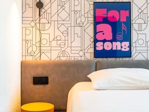 Una habitación moderna con un papel tapiz de diseño geométrico, una cama con almohada blanca, cabecero gris y una mesita de noche amarilla.