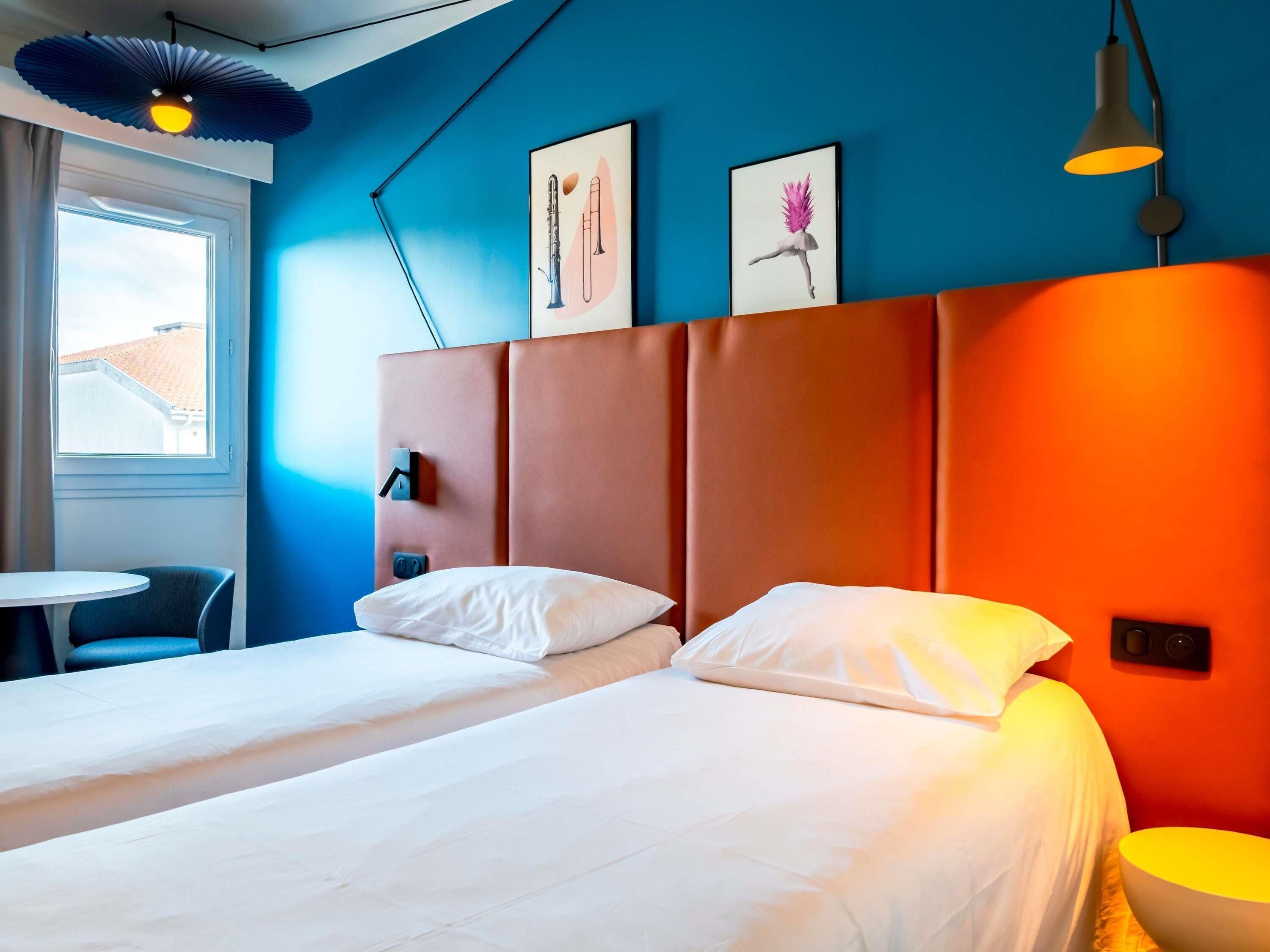 A hotel room with two beds, a small table, a chair, and artwork on the wall.