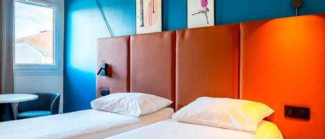 A hotel room with two beds, a small table, a chair, and artwork on the wall.