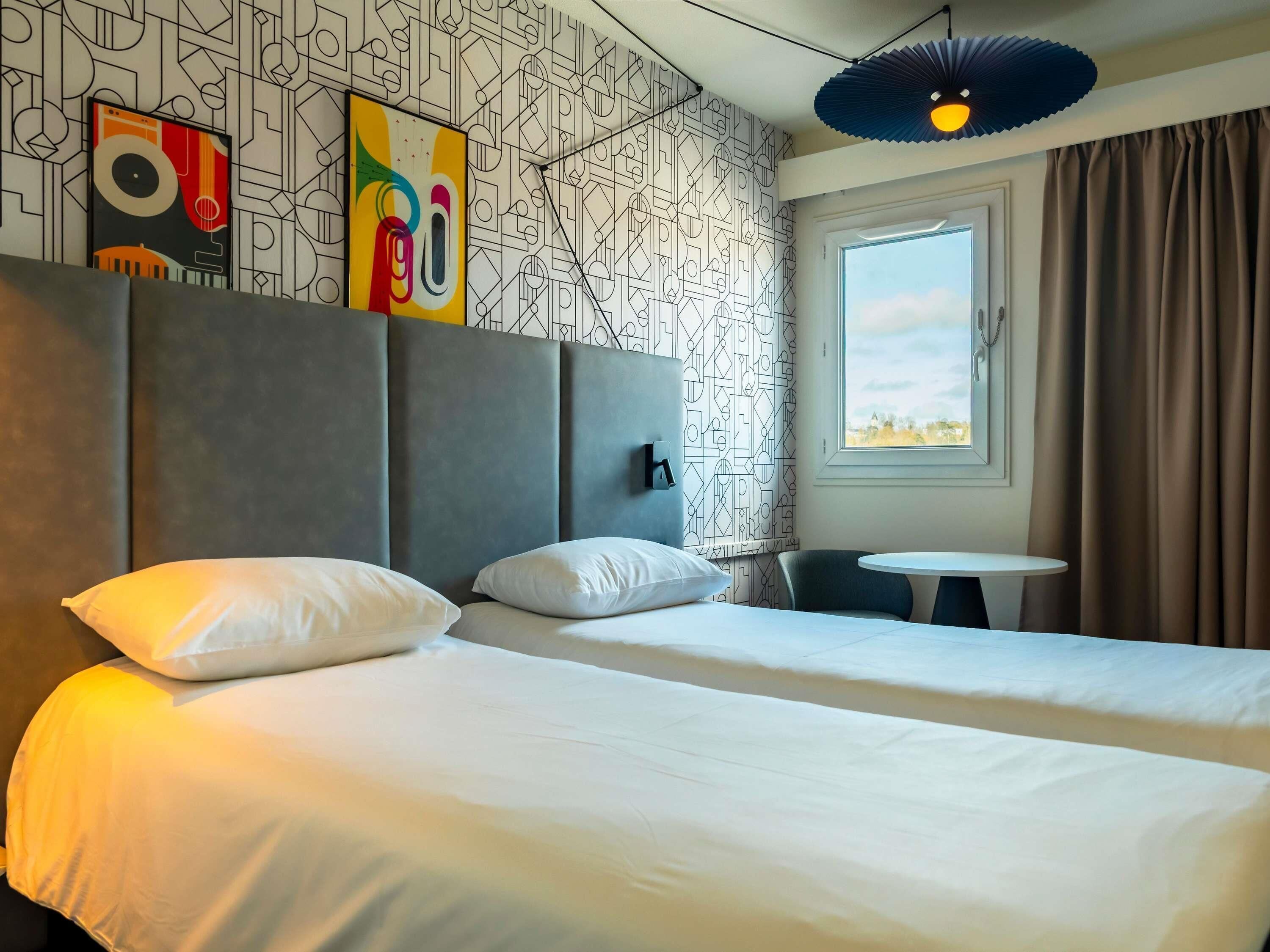 A hotel room with two beds, a small table, a window with curtains, and geometric patterned wallpaper.