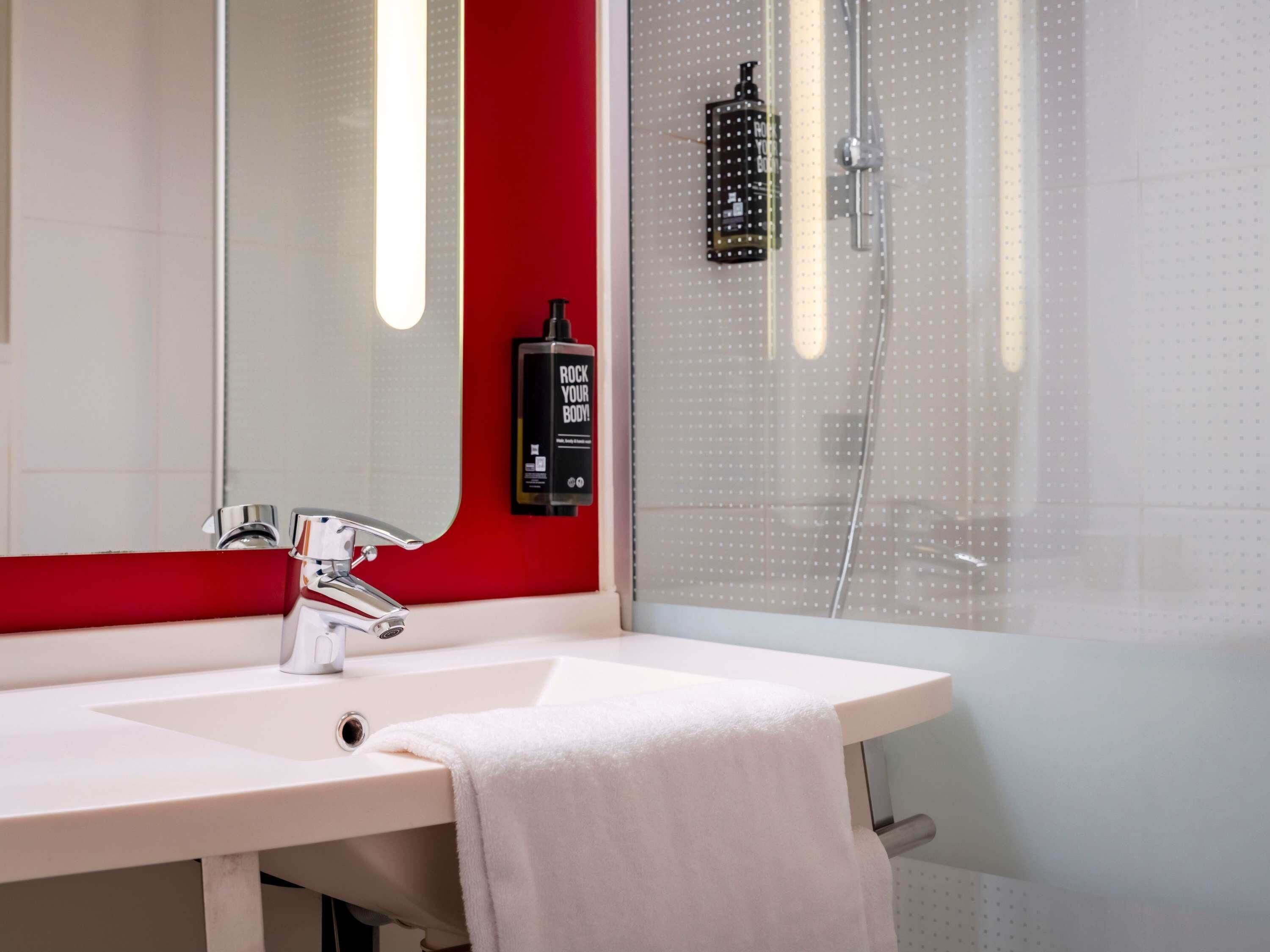 A bathroom with a white sink, a mirror, a red accent wall, a shower with a glass door, and a soap dispenser.