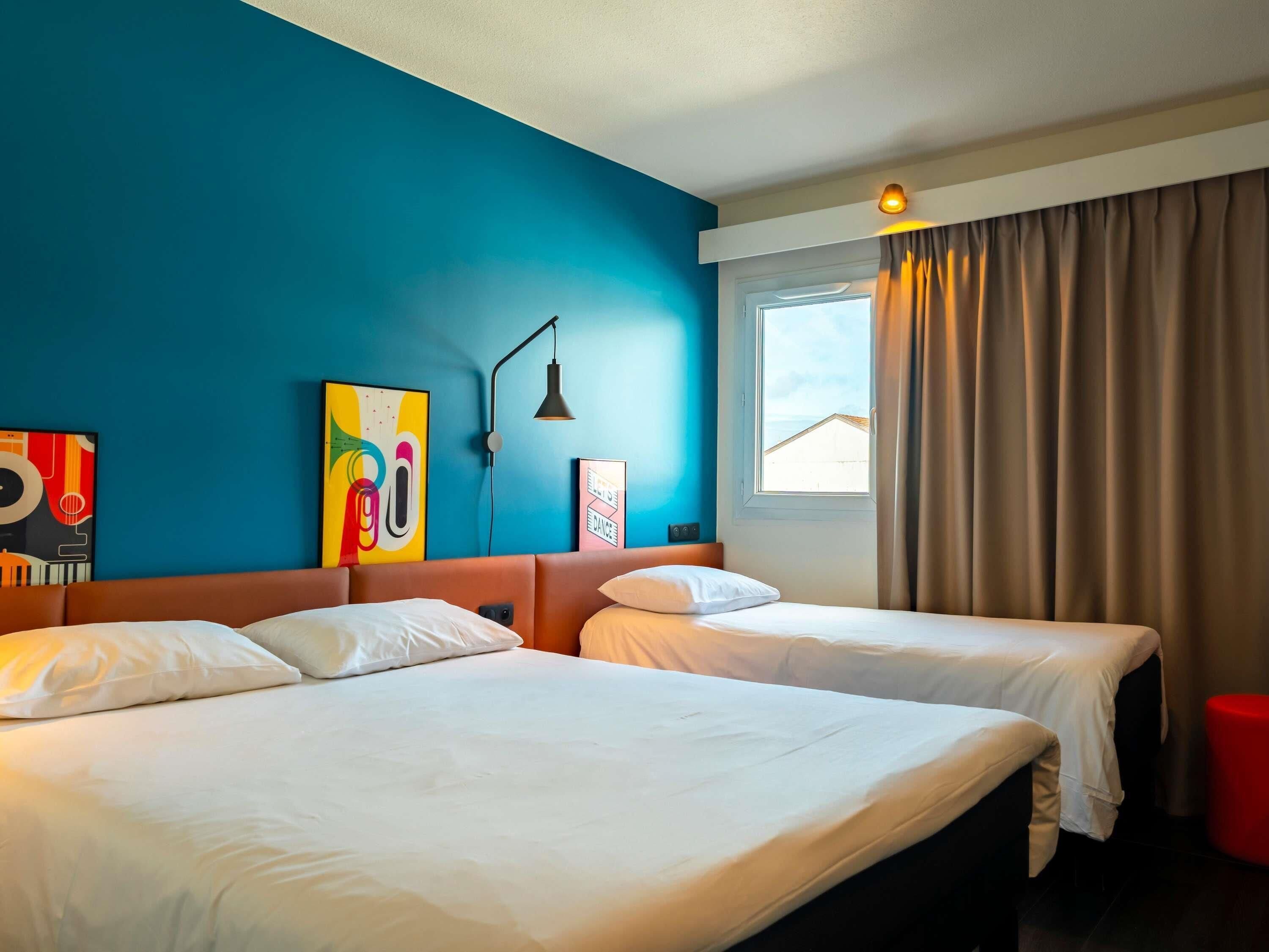 Una habitación de hotel con dos camas, una ventana con cortinas, un cuadro abstracto en la pared y una lámpara.