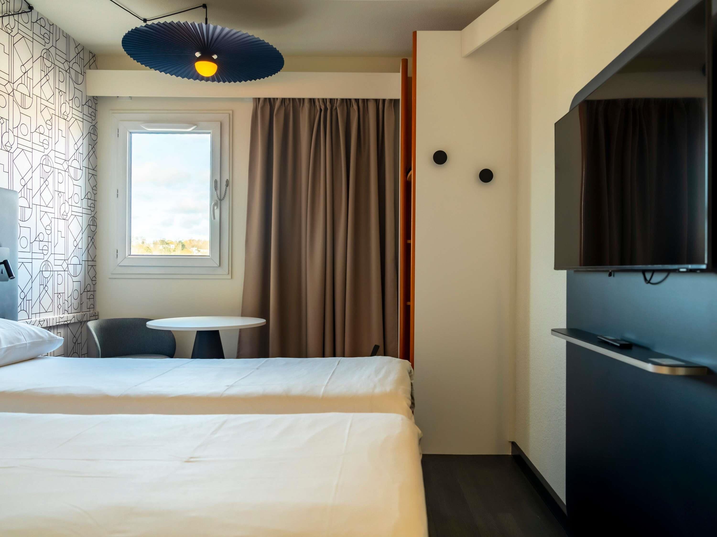 Une chambre d’hôtel avec un lit, une petite table, une chaise, une télévision et une fenêtre avec des rideaux.