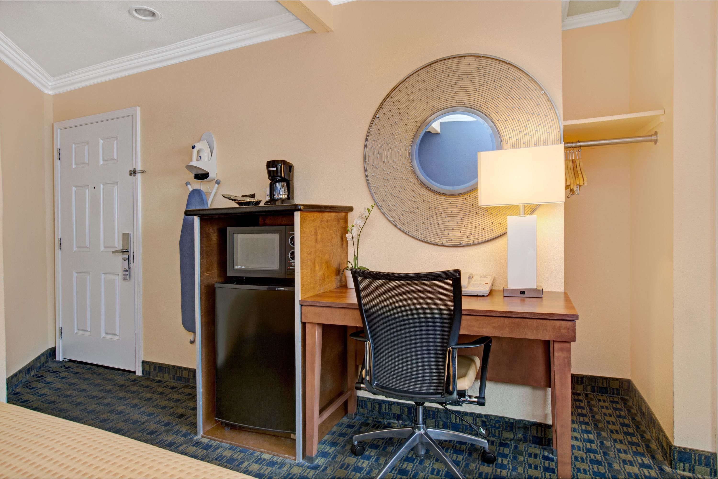 A hotel room with a desk, chair, microwave, coffee maker, and a round mirror on the wall.