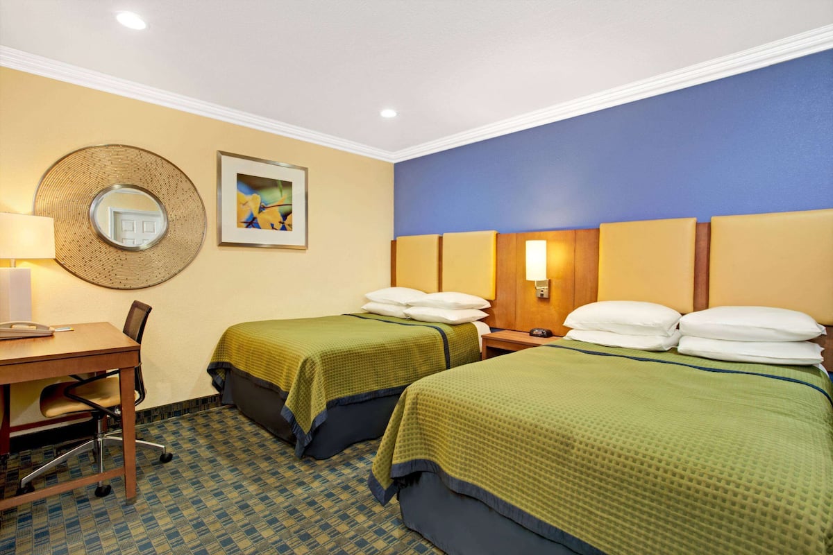 Une chambre d’hôtel avec deux lits, un bureau, une chaise, une lampe, un miroir et un tableau encadré au mur.