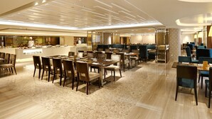 A spacious hotel dining area with multiple tables and chairs, a central bar, and a kitchen visible in the background. - Prudential Hotel (Kowloon)